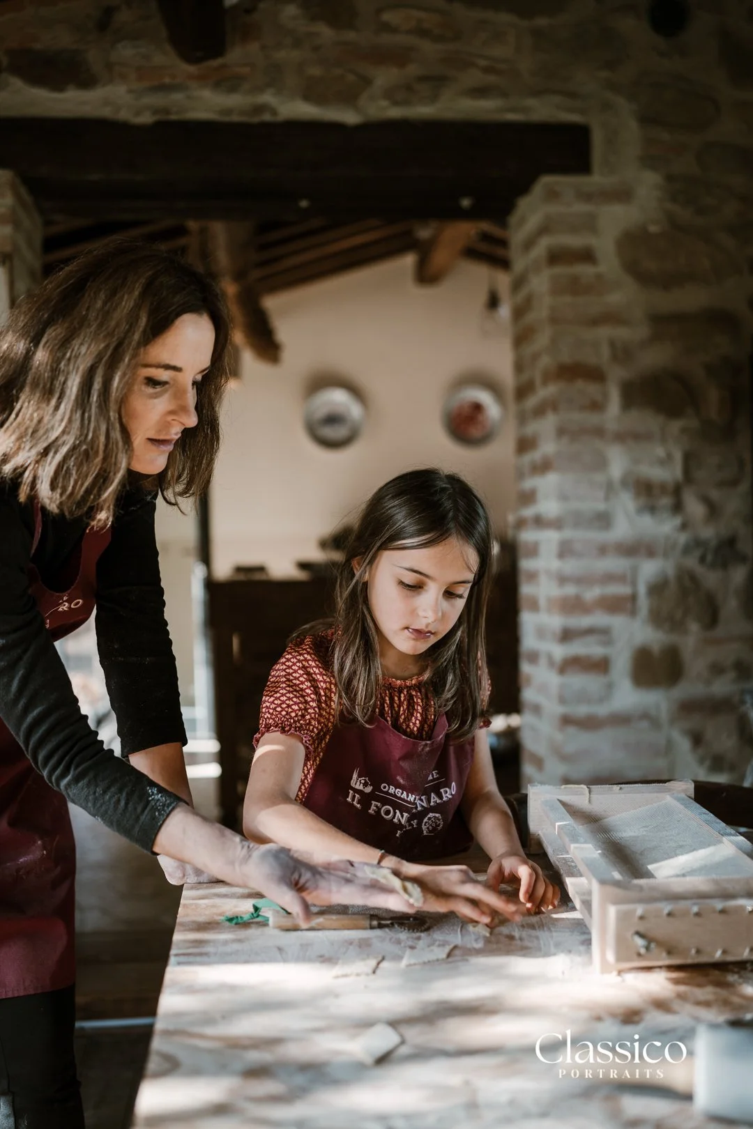 A woman and a young girl working together on a craft project at a wooden table in a rustic, brick-walled room with decorative plates on the wall.