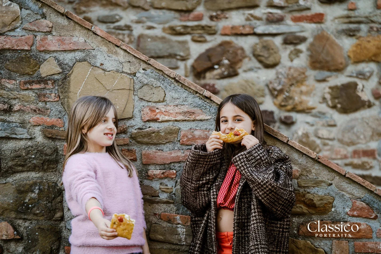 Two young girls standing outdoors against a stone and brick wall, holding slices of pizza, one girl is eating her slice and the other girl is offering her slice.