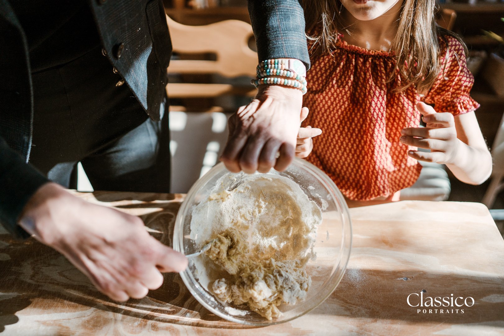 An adult and a young girl are baking together; the adult is mixing dough in a glass bowl, and the girl is holding her hands near the bowl, watching the process. The scene is set on a wooden table in a cozy, well-lit kitchen.
