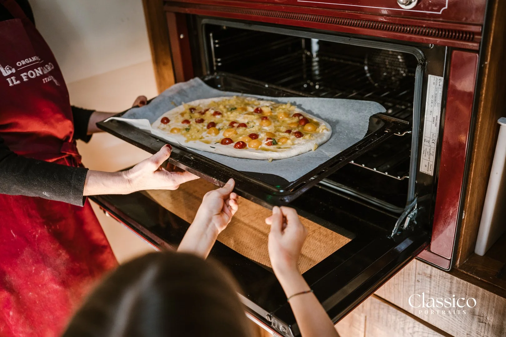 Person removing a pizza with cherry tomatoes from an oven while another person points at it.