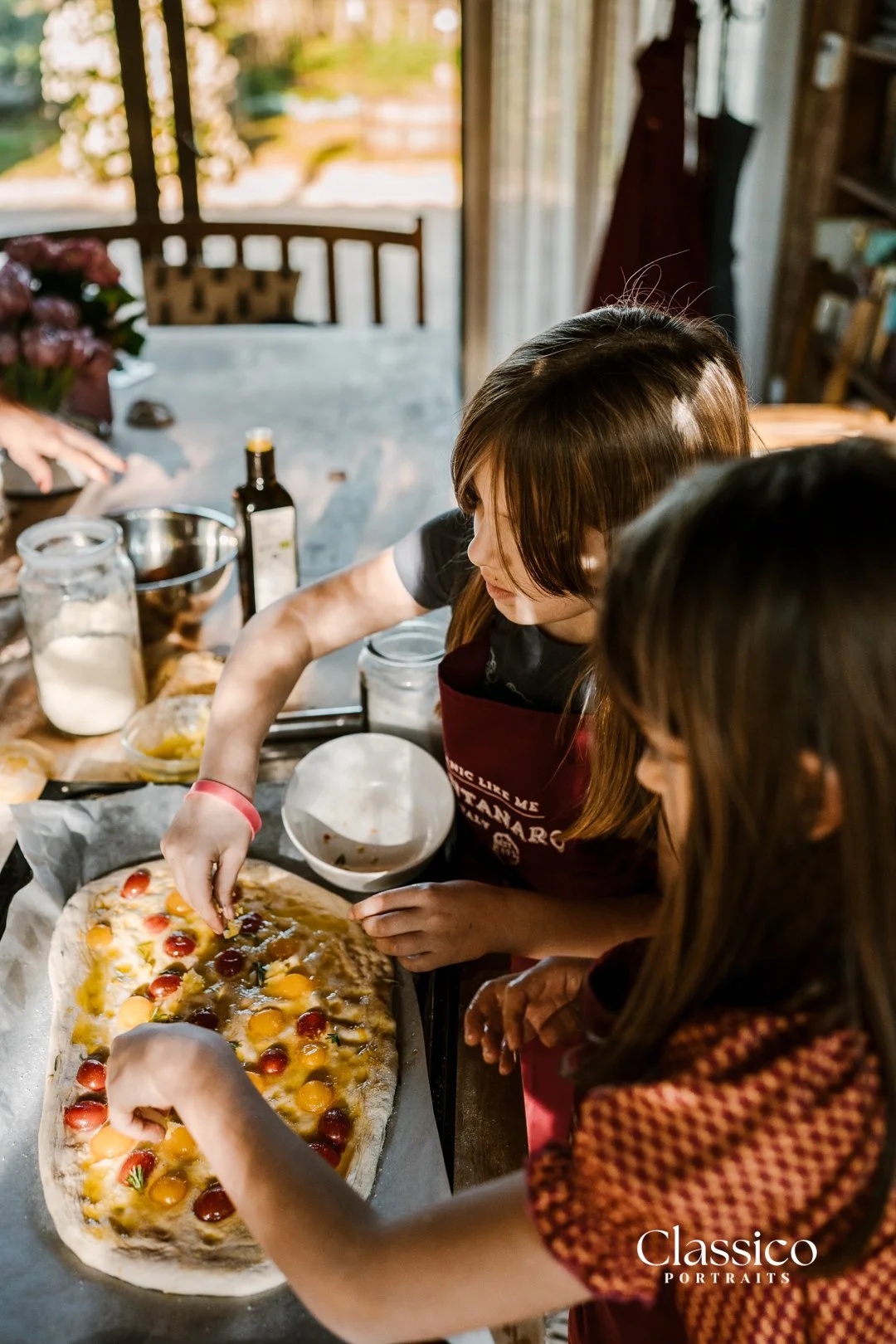Two women are making a homemade pizza in a rustic kitchen, adding toppings to the dough on a baking sheet.