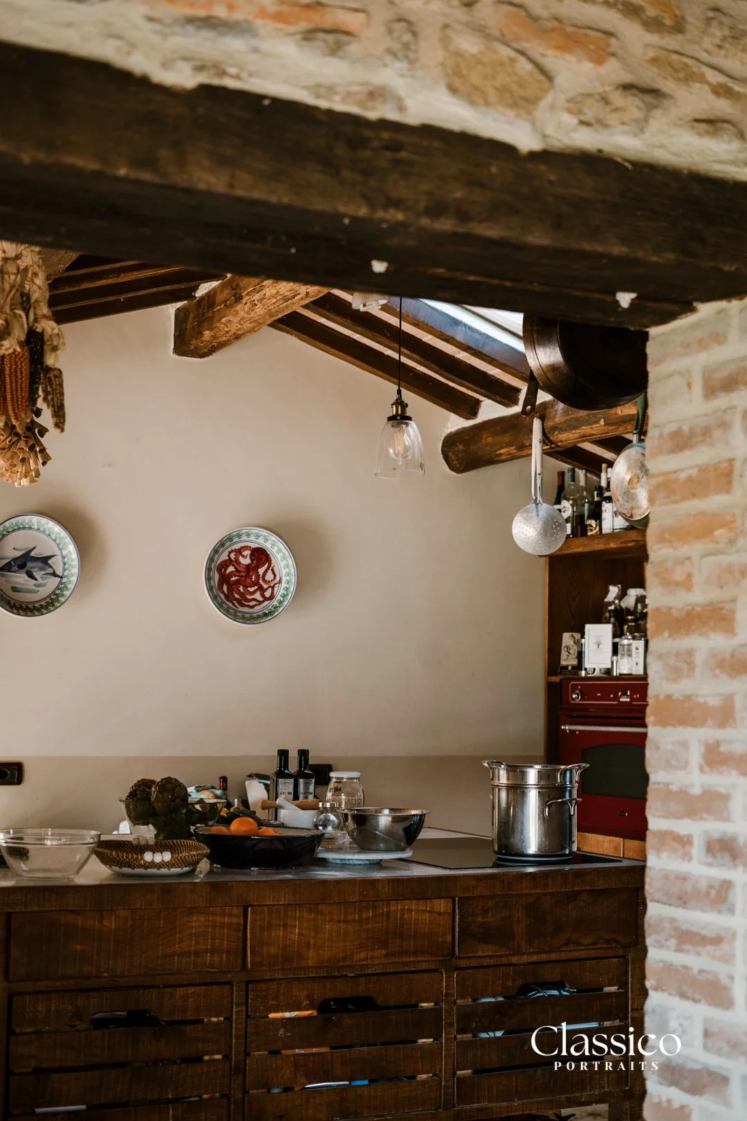 A rustic kitchen with wooden beams, brick walls, hanging kitchen utensils, and a countertop with various bowls, bottles, and cooking pots.