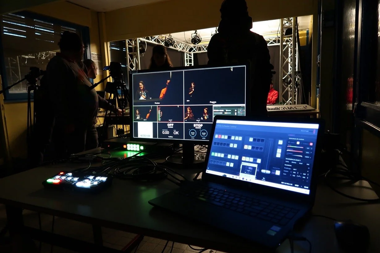 Sala de contrôle technique, avec deux écrans, un ordinateur portable, des consoles et des câbles, en arrière-plan des personnes en train de préparer une scène avec un éclairage professionnel.