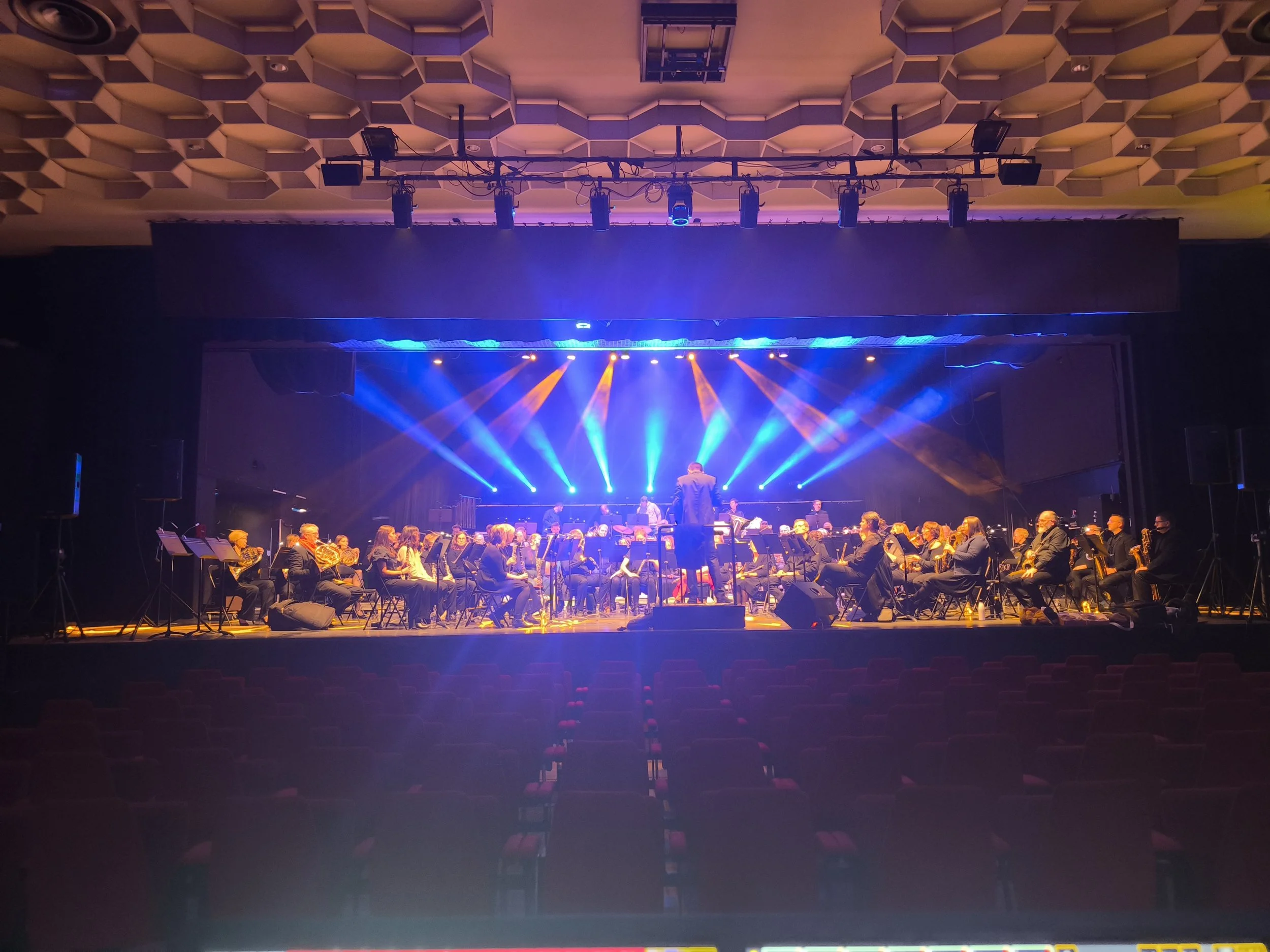 Orchestre symphonique sur scène, dirigé par un chef d'orchestre, avec des éclairages colorés en bleu et orange, et un auditorium vide en premier plan.
