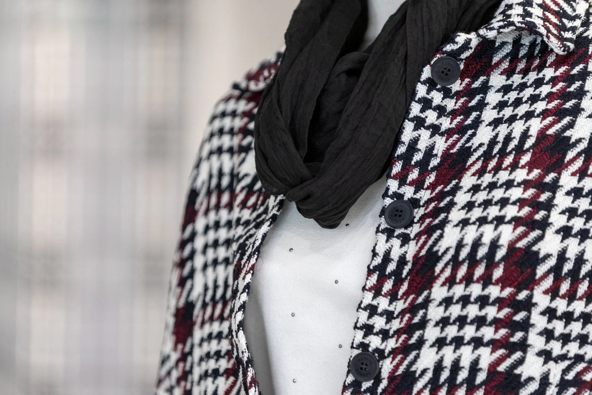 Nahaufnahme einer Person, die einen schwarzen Schal trägt, ein kariertes Jacke in schwarz, weiß und rot, mit großen schwarzen Knöpfen, und ein weißes Shirt darunter, mit kleinen Schmucksteinen.