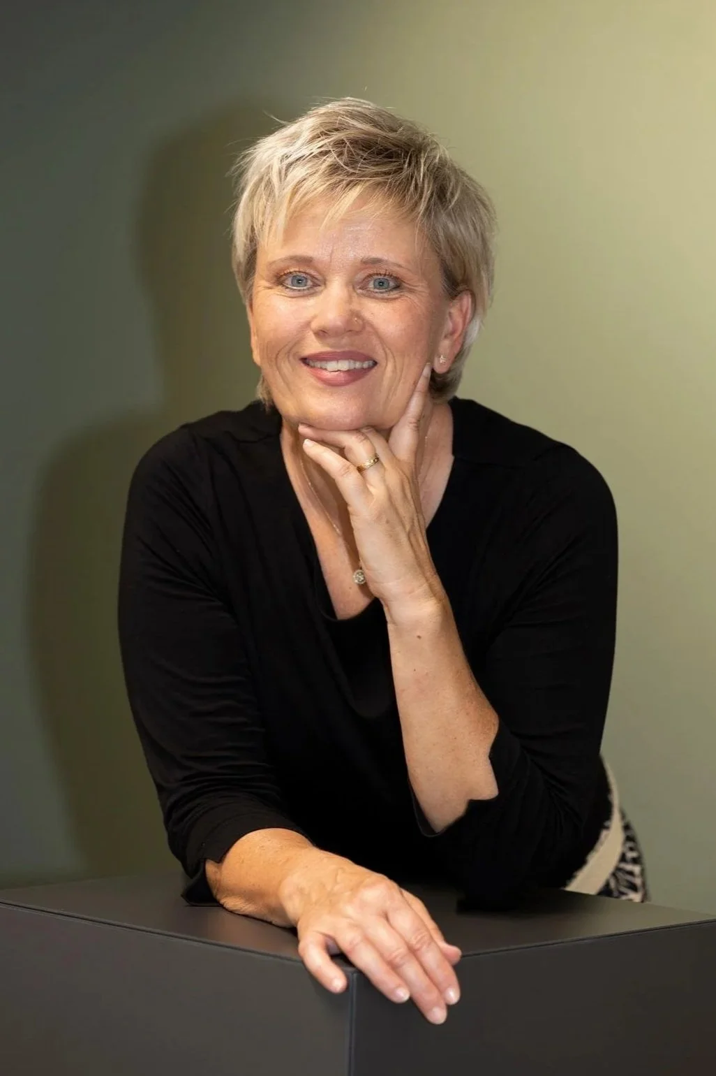 Frau mit kurzer, grauer Frisur, in schwarzer Kleidung, lächelnd und mit der Hand unter dem Kinn am Tisch sitzend, vor grünem Hintergrund.