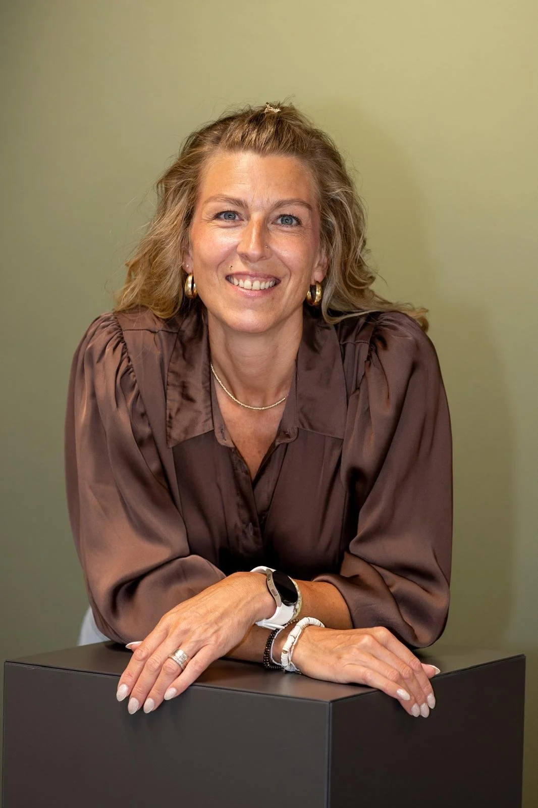 Eine Frau mit blonden, lockigen Haaren, die ein braunes Seidenhemd trägt, sitzt lächelnd vor einem grünen Hintergrund.