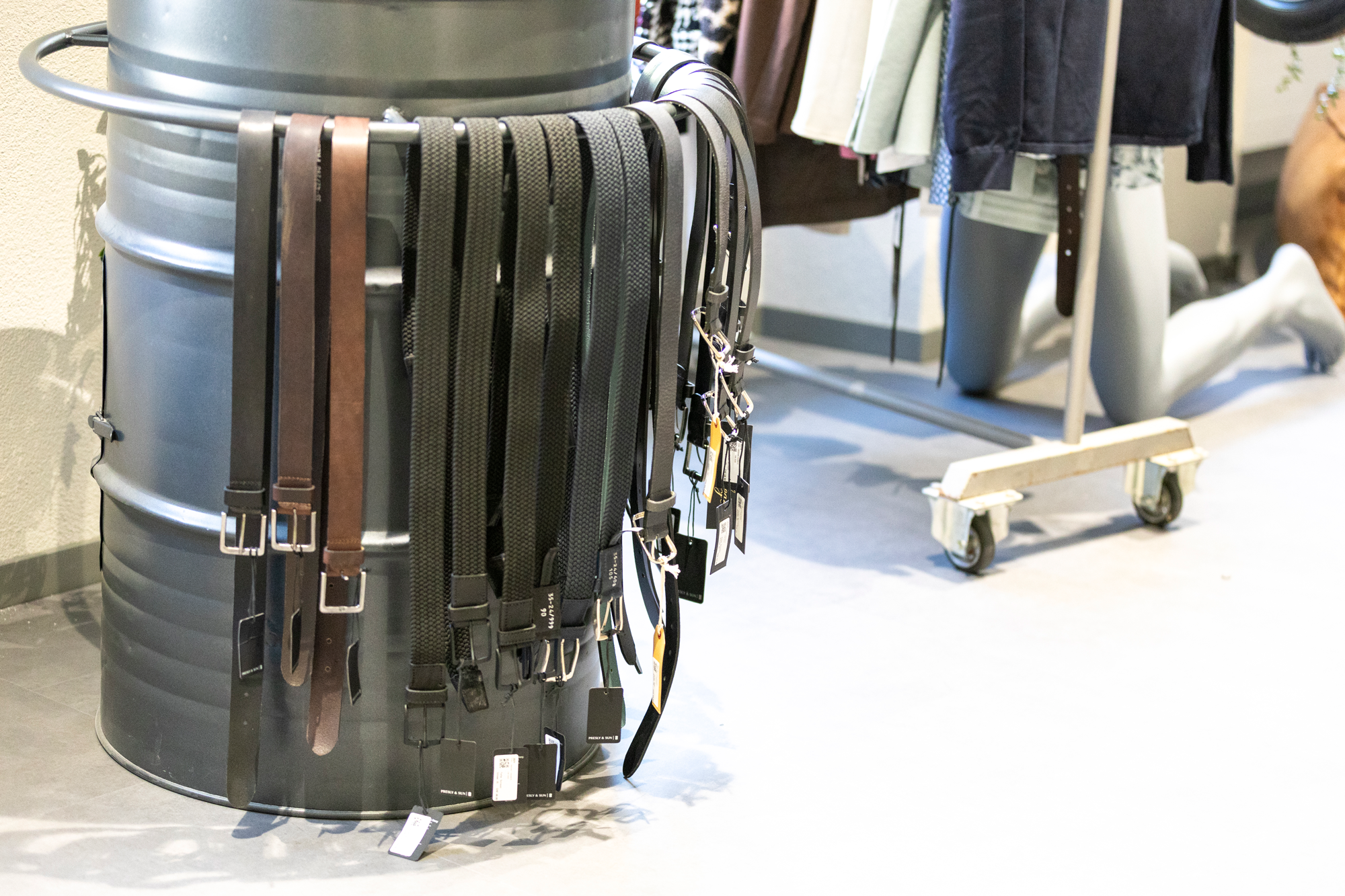 Mehrere Gürtel in verschiedenen Farben hängen an einer runden Metallstand, im Hintergrund Kleidung auf einem Kleiderständer.