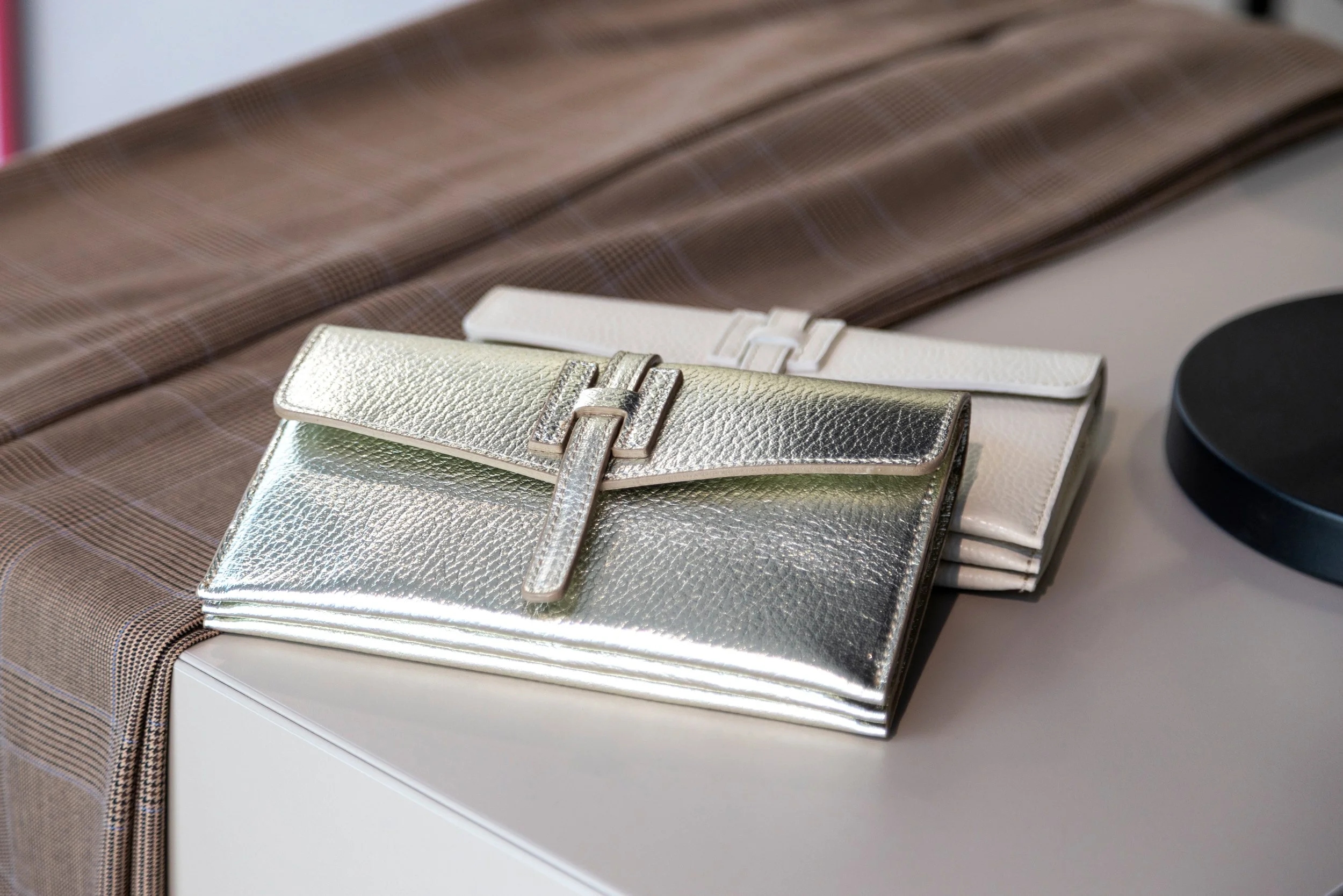 Drei Taschen in Metallic-, Creme- und Silberfarben liegen auf einem weißen Möbelstück. Im Hintergrund ist ein brauner Stoff mit Karo-Design. Rechts befindet sich eine schwarze runde Gegenstand.