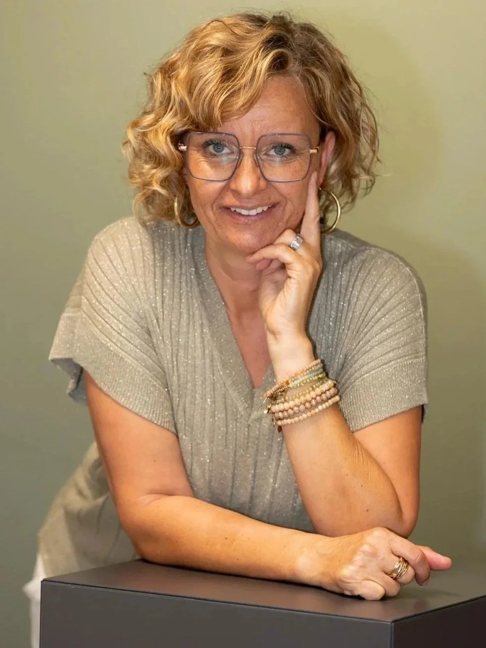 Eine lächelnde Frau mit lockigem, schulterlangem Haar, trägende Brille, trägt eine beige, glitzernde Bluse und mehrere Armbänder. Sie ist mit einer Hand an ihrem Kiefer und der anderen auf einem schwarzen Tisch. Der Hintergrund ist neutral.
