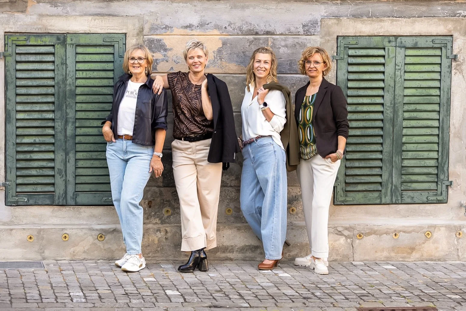 Vier Frauen stehen vor einer alten Hauswand mit grünen Fenstergittern, sie lachen und posieren für die Kamera.