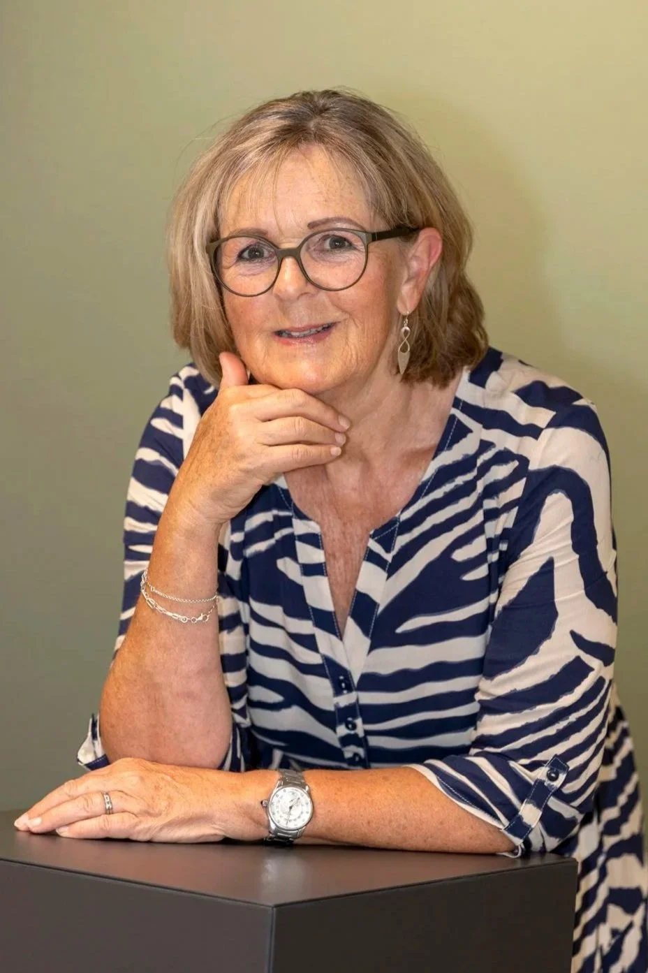 Porträt einer älteren Frau mit Brille, kurzer hellbrauner Haar, Musterbluse in Navy und Weiß, sitzt an einem Tisch, lächelnd, mit Hand am Kinn, trägt eine Armbanduhr und Silberohrringe.