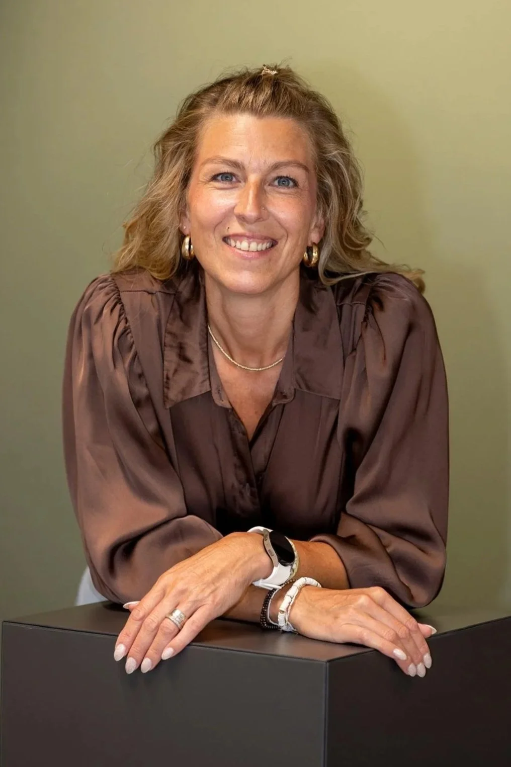 Eine lachende Frau mit lockigem, braunem Haar, goldenen Ohrringen, brauner Bluse, sitzend an einem Tisch vor einem grünen Hintergrund.