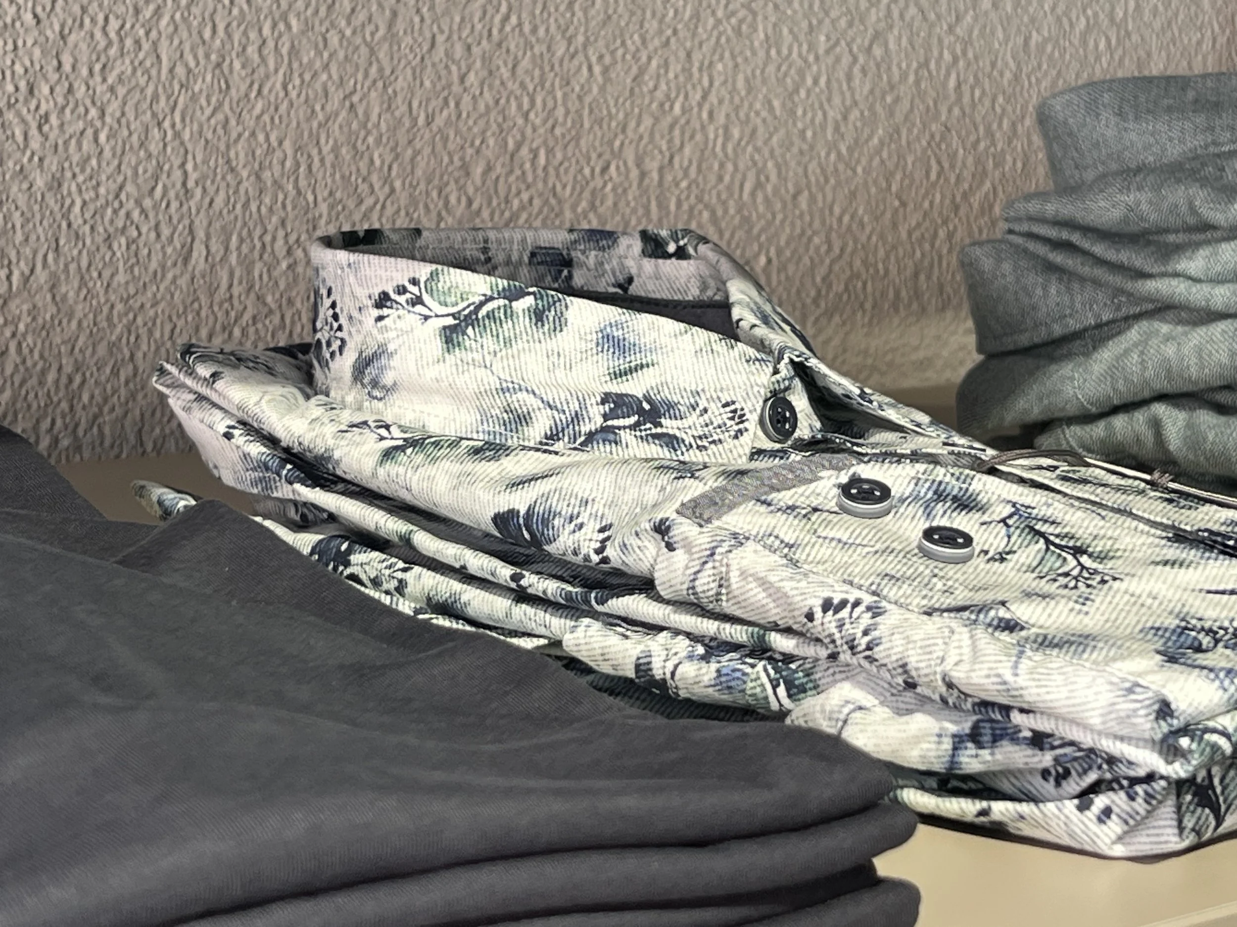 Ein Bild zeigt die unteren Körperteile einer Person, die auf einem grauen Teppich sitzt. Die Person trägt eine Hose mit einem schwarzen, weißen, grünen und blauen Blumenmuster. Es sind noch andere Kleidungsstücke und Gegenstände im Bild sichtbar.