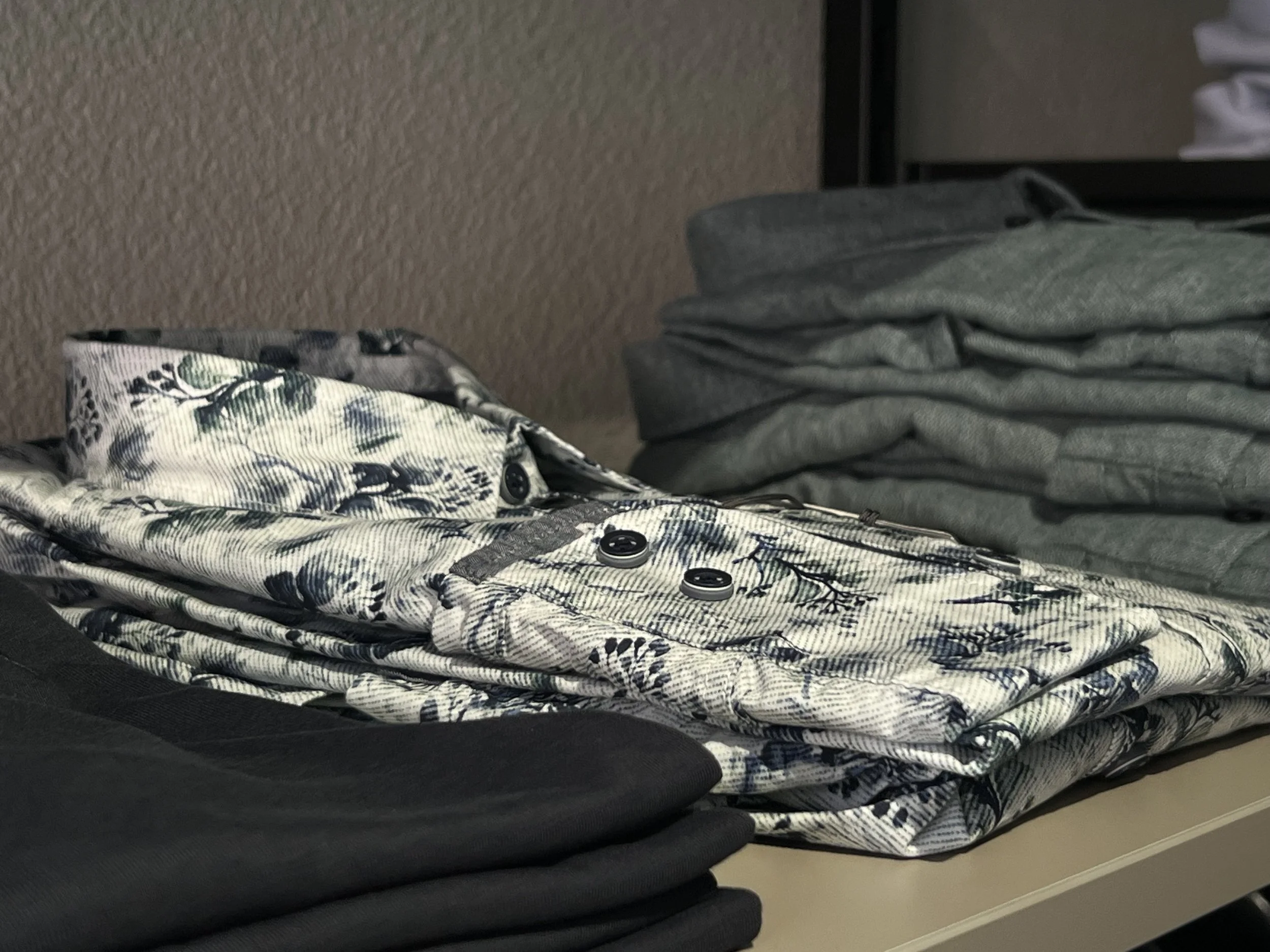 Ein Stapel Kleidung besteht aus einem gemusterten Hemd, einer grauen Jacke, grauen Hosen und weiteren Kleidungsstücken auf einer Ablage im Büro.