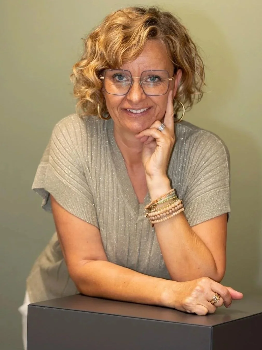 Eine Frau mit lockigem, blondem Haar trägt Brille, ein silbernes Shirt und mehrere Armbänder, lehnt mit einem Arm auf einer schwarzen Oberfläche und lächelt in die Kamera.