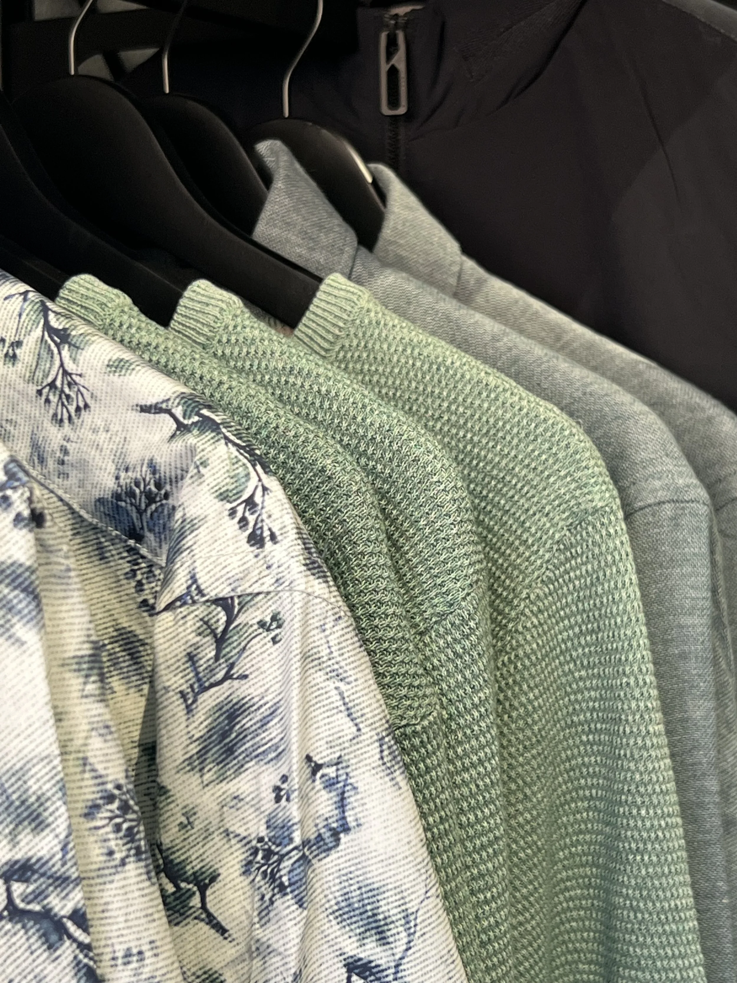 Nahaufnahme von mehreren Kleidungsstücken auf Kleiderbügel in einem Schrank, inklusive Bluse mit Blumenmuster, grüner Hemd und graues Kleid.