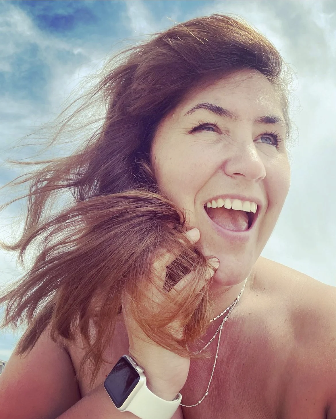 A woman with brown hair smiling and holding her hair, wearing a smartwatch and a necklace, against a blue sky background.