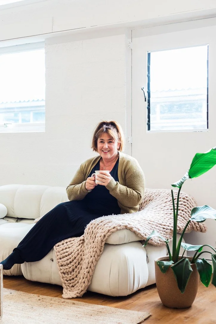A woman sitting on a white sofa in a brightly lit room, holding a coffee mug and smiling, with a large green plant nearby.