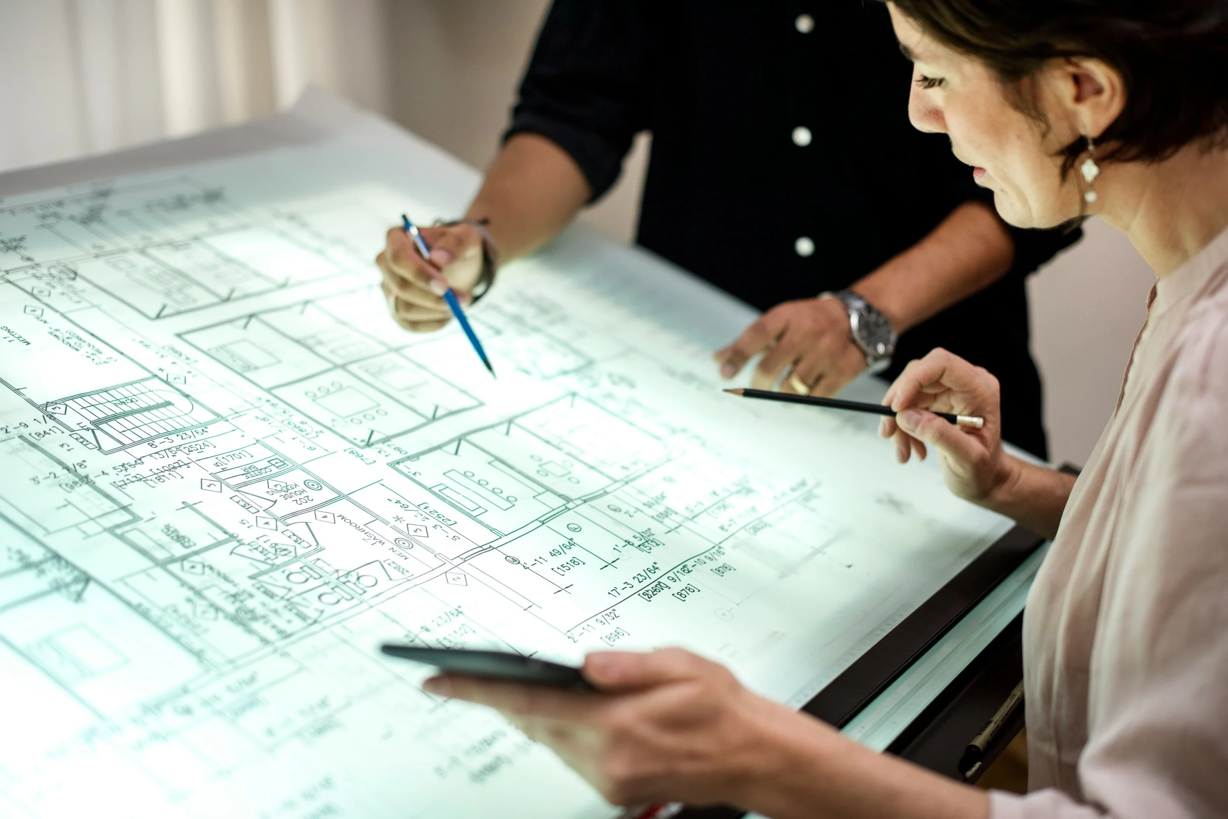 Twee mensen bekijken en bespreken een bouwplan of architectuurontwerp op een grote verlichte tafel, met documenten en een smartphone in de hand.
