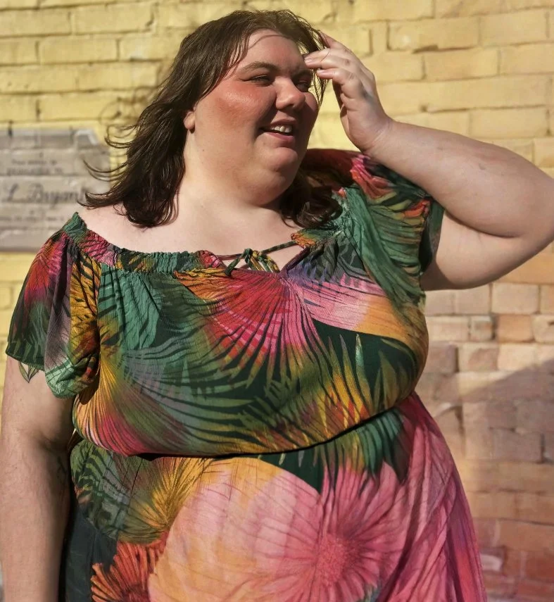A smiling woman with shoulder-length brown hair wearing a colorful tropical patterned dress, standing in front of a brick wall.