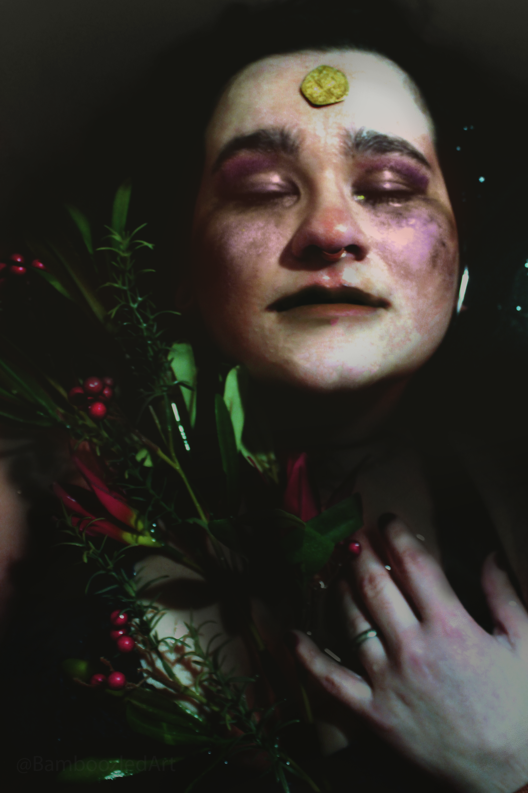 A person with makeup, closed eyes, and a penny on their forehead, submerged in water with plants and berries around them.