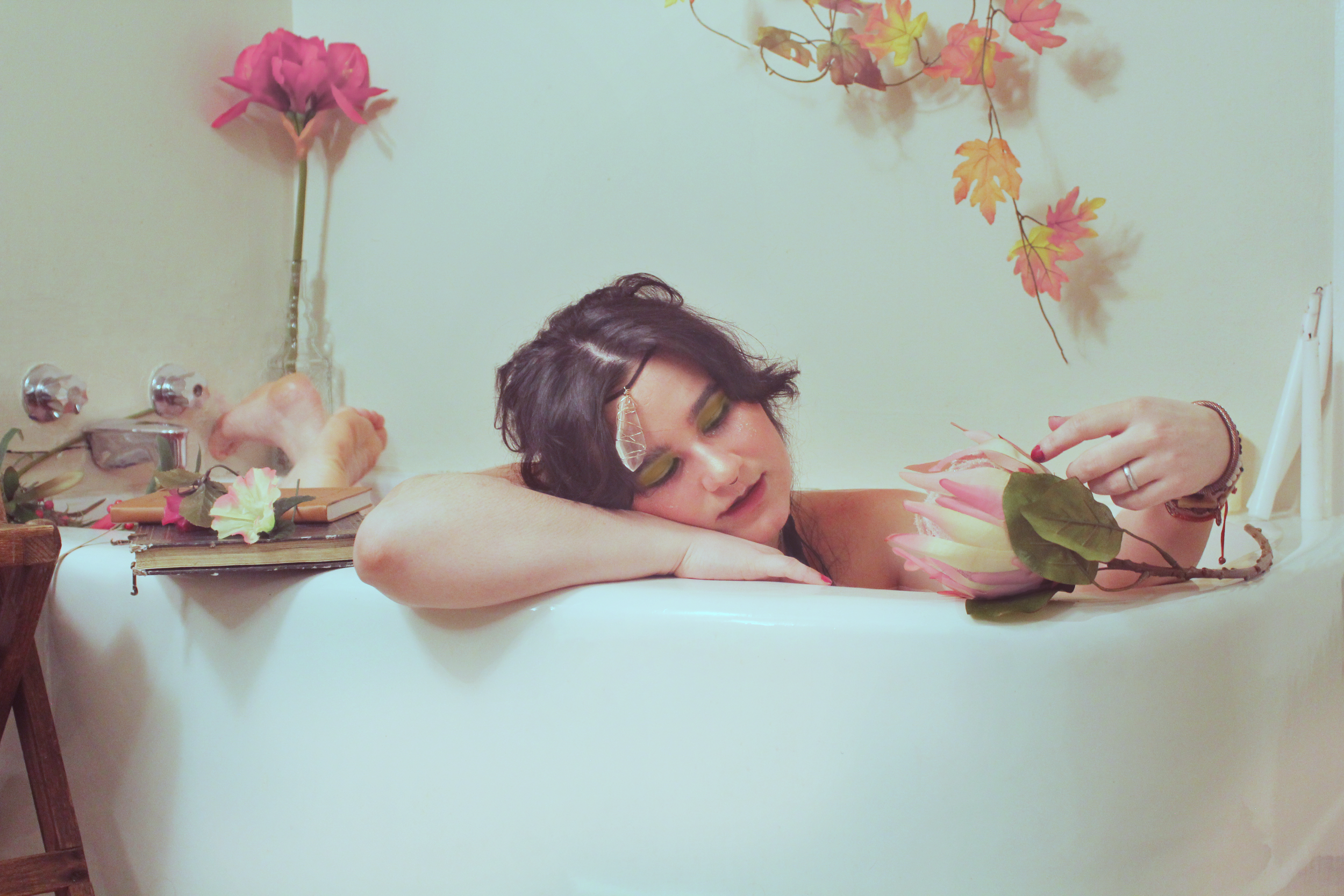 Woman with black hair and green eyeshadow relaxing in a bathtub, holding a pink flower, with pink and yellow leaves and a pink flower in the background.