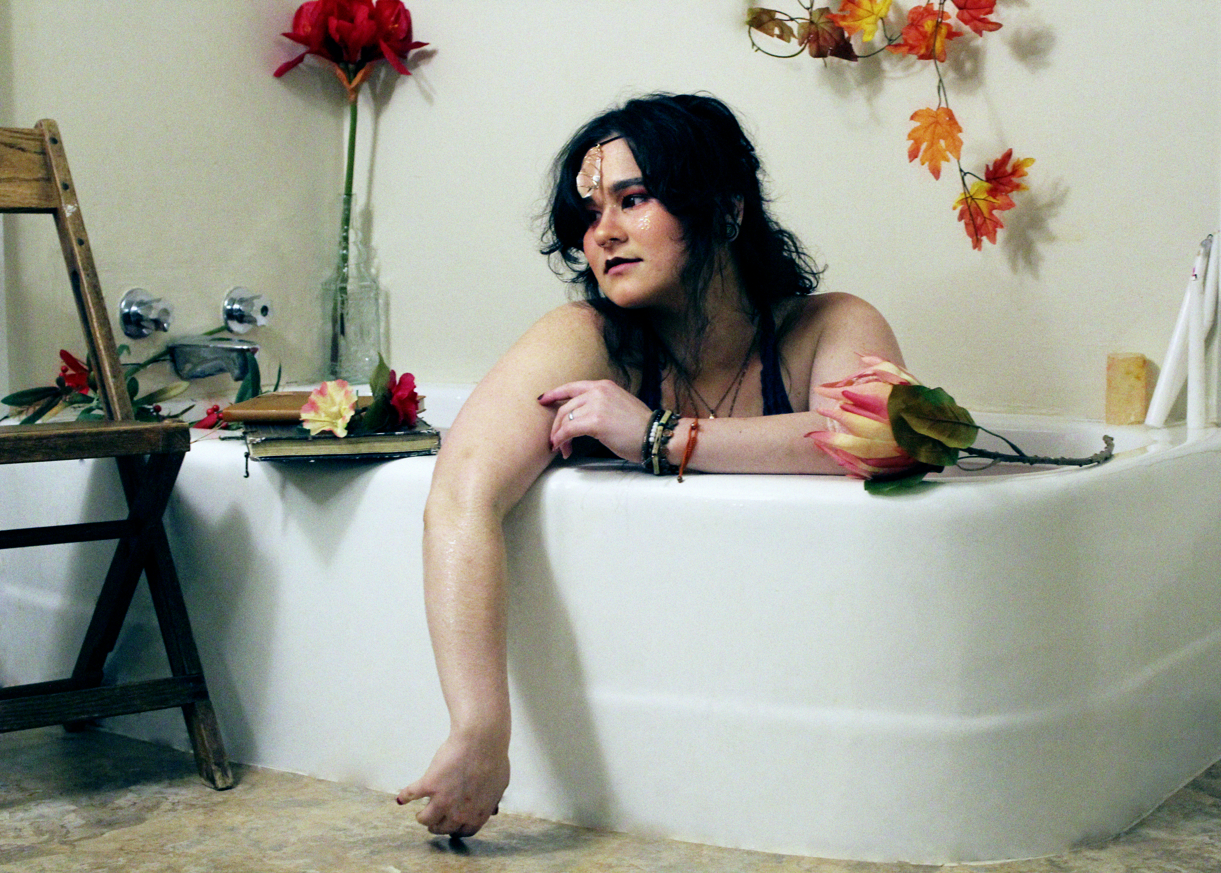 Woman with dark hair sitting in a bathtub with her arm hanging over the edge, surrounded by flowers and leaves, with a wooden chair, a mirror, and a beige wall in the background.