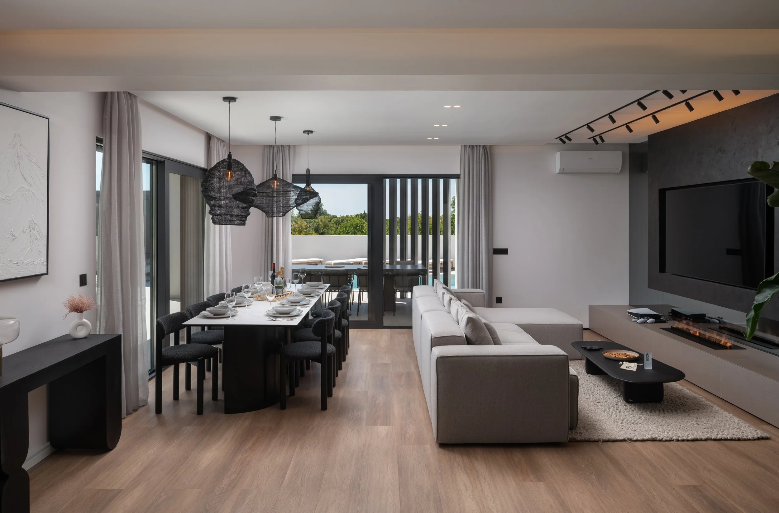 Modern living room with dining area, white sofa, black coffee table, mounted flat-screen TV, and glass sliding door leading to outdoor balcony with green trees.