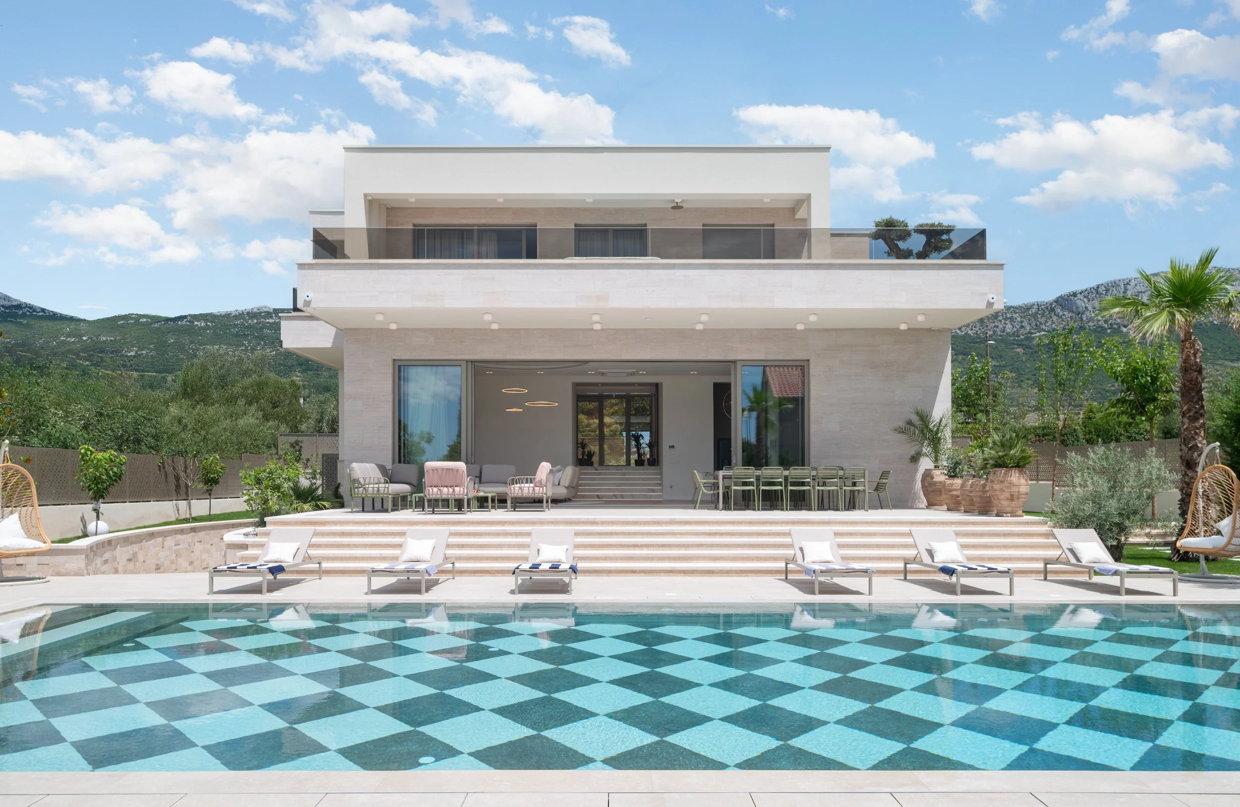 Luxury modern house with large swimming pool, lounge chairs, outdoor seating area, potted plants, and scenic mountain view in the background under a blue sky with some clouds.