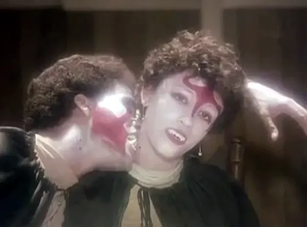Two individuals with red face paint, fangs, and black capes, with one looking past the camera and the other biting the neck of the first.