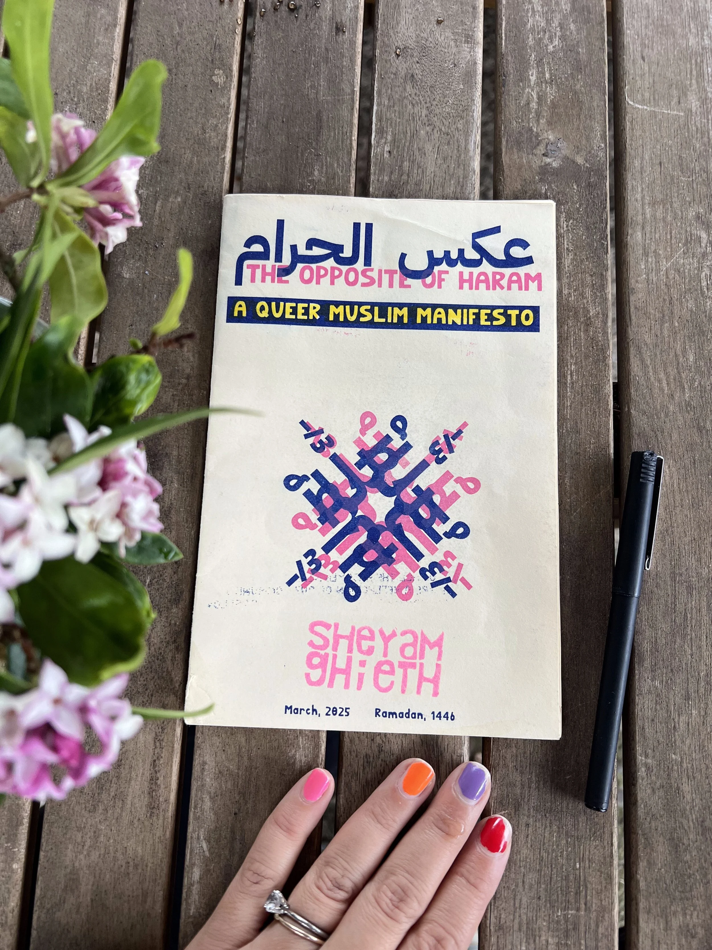 Graphic of geometric pink and blue artwork against a white background in the center of a zine entitled "The Opposite of Haram: A Queer Muslim Manifesto" by Sheyam Ghieth. The zine is resting on a plank table, with flowers on the left.