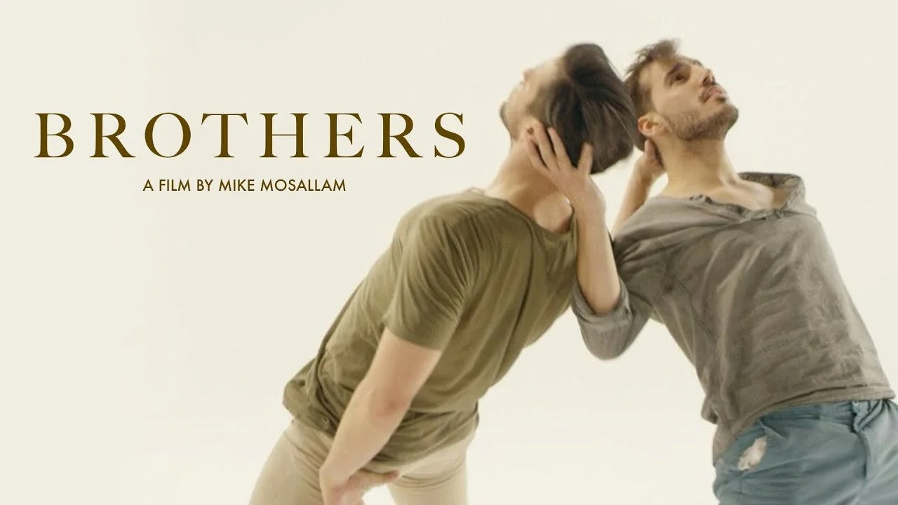 Two men with messy hair and casual clothing are holding the back of each other's heads as they each lean back. The background is plain and the text on the image reads "Brothers" and "A film by Mike Mosallam."