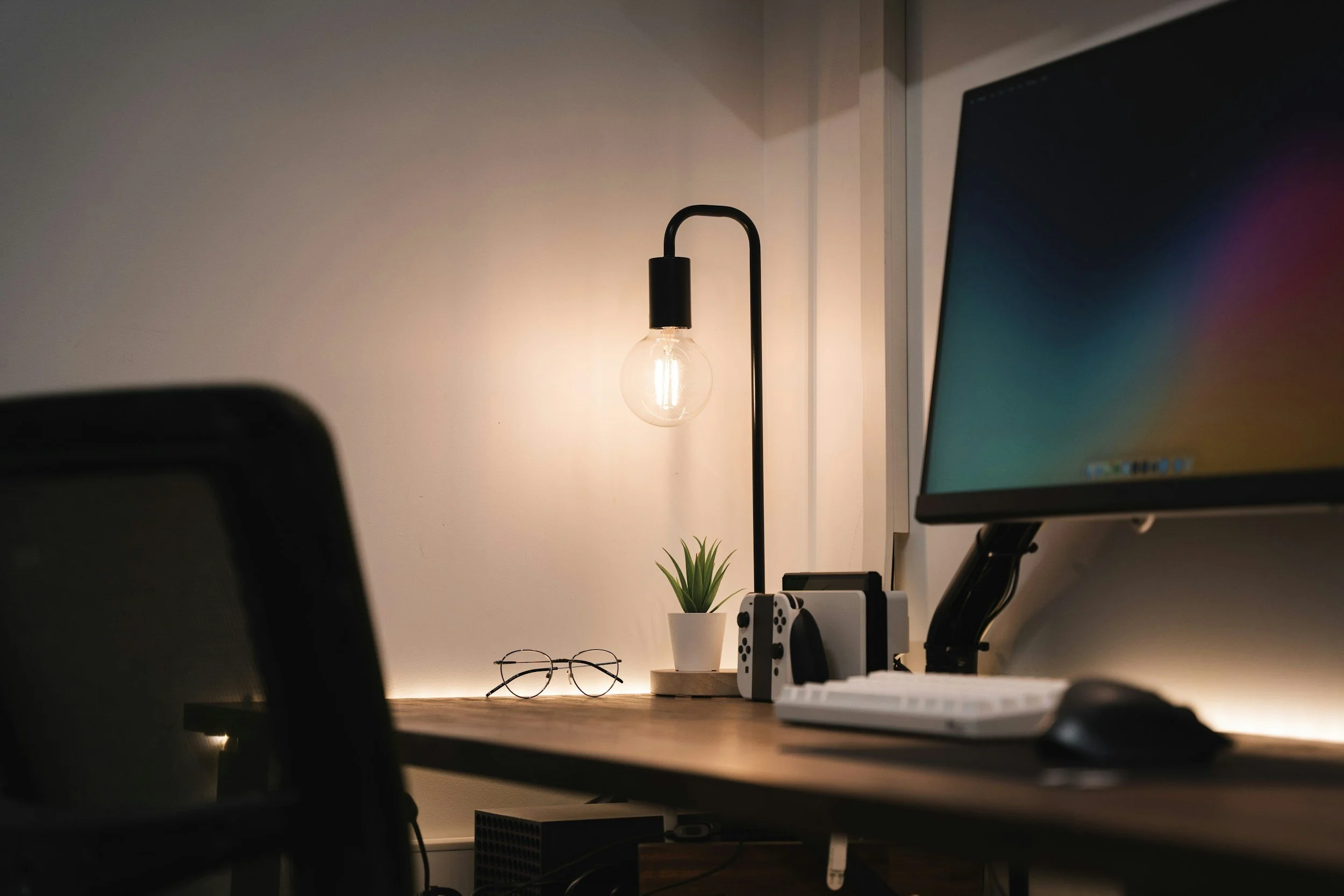 A workspace with a modern desk lamp, a small potted plant, a pair of glasses, and a computer monitor on a wooden desk.