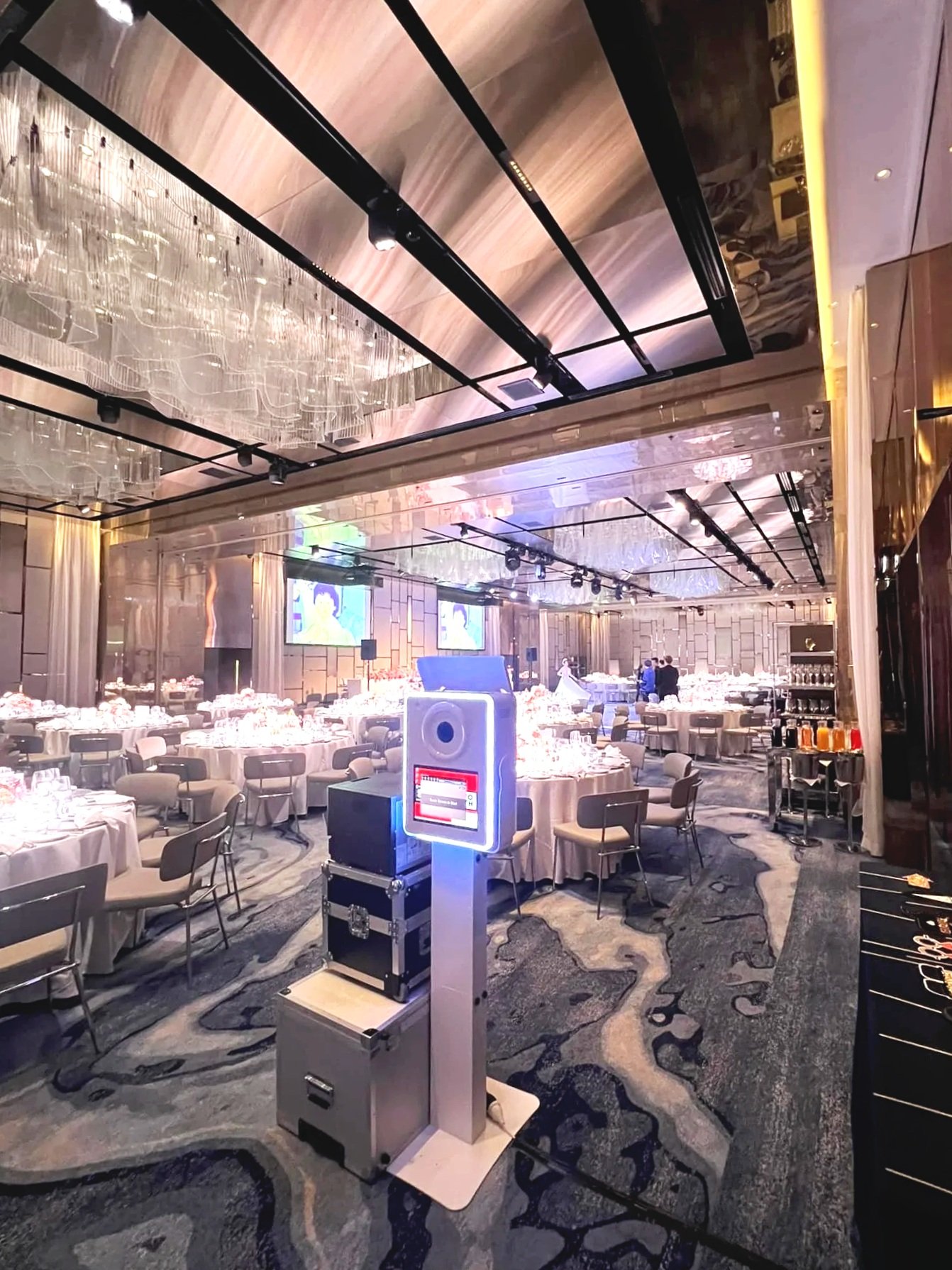 A banquet hall with round tables covered in white tablecloths and set with glassware and silverware. The hall has a modern design with a reflective ceiling, black framing, and large screens on the wall. A photo booth with a camera and lighting equipment is positioned in the foreground.