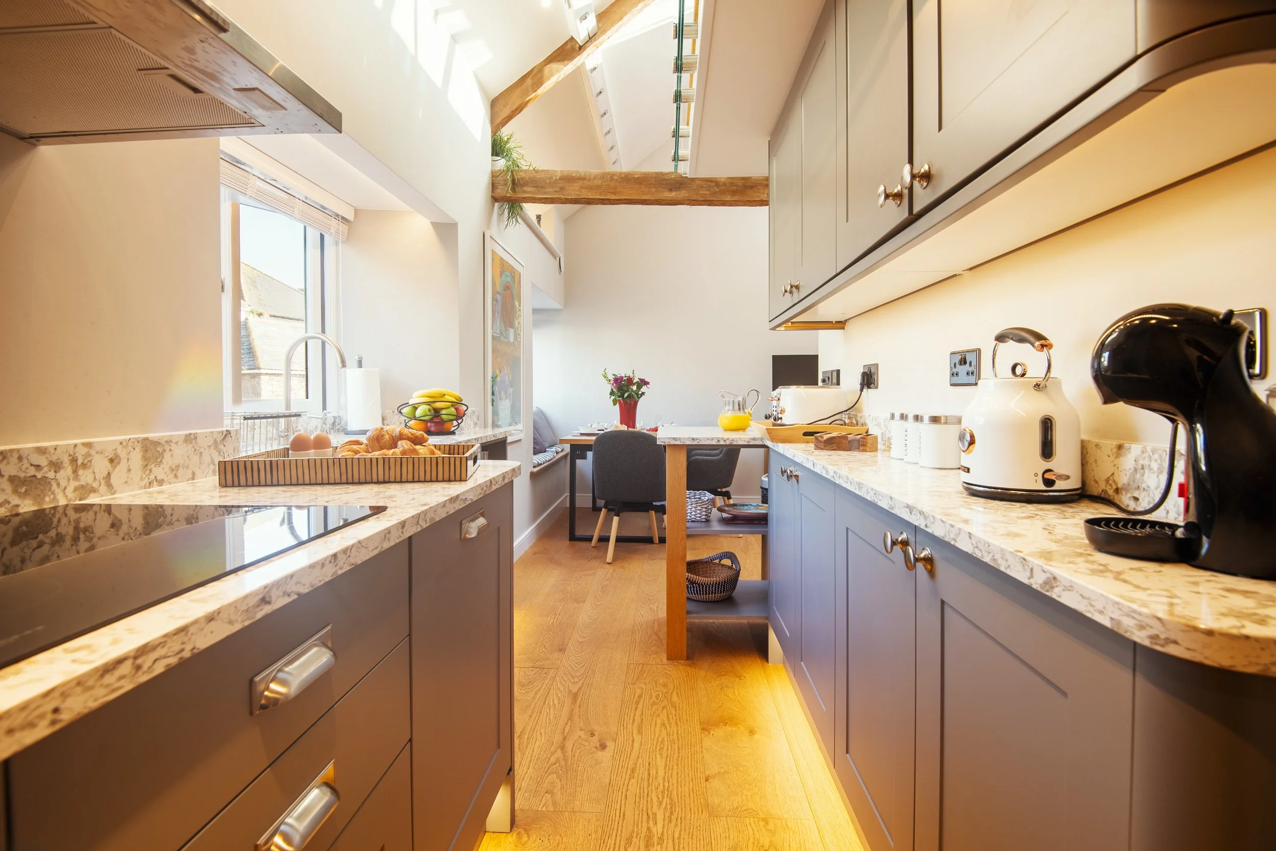 Bright kitchen with beige marble countertops, gray cabinets, wooden beams, a window with sunlight, and a small dining area with a table, chair, and flowers.