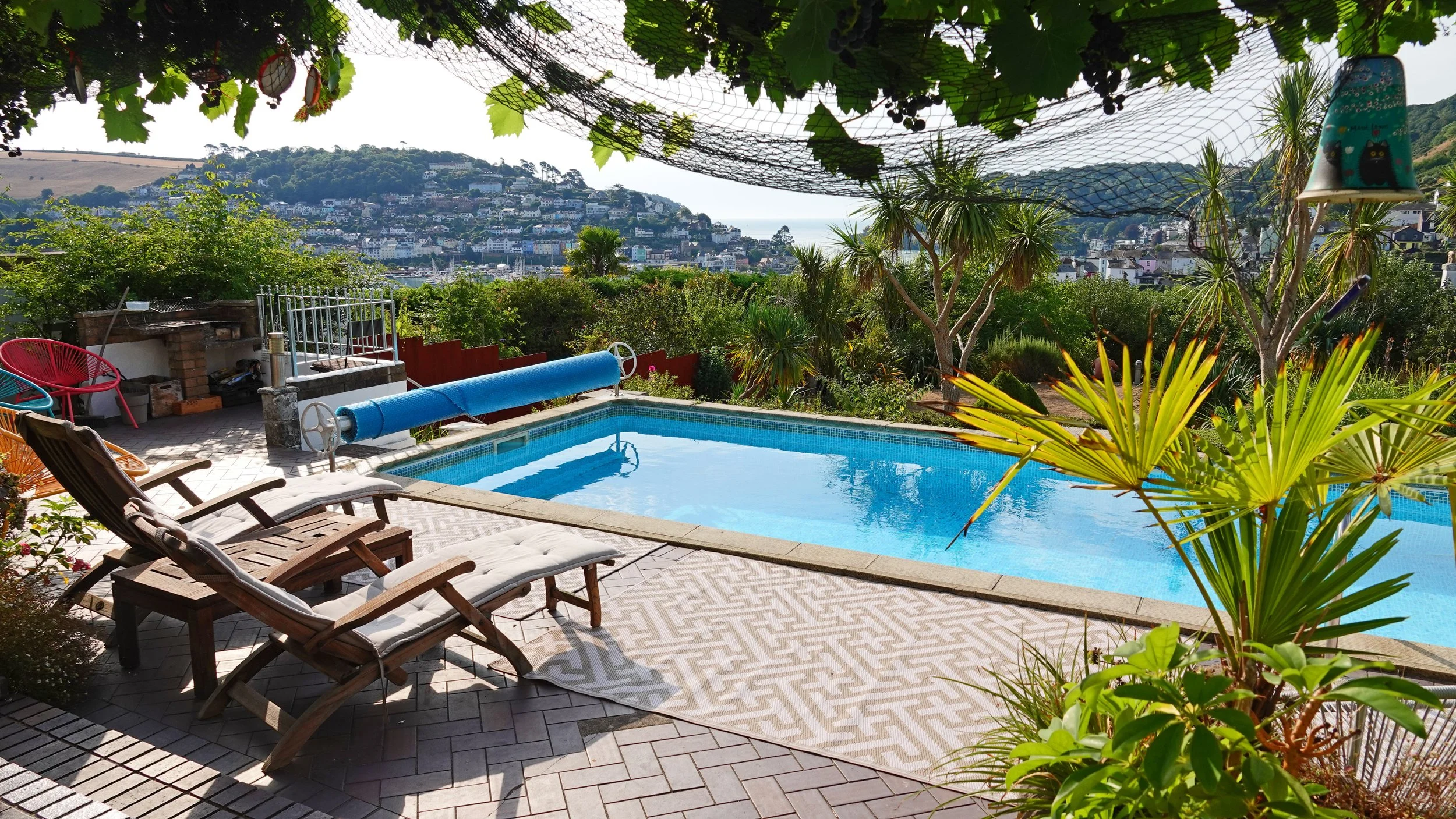 A backyard with a swimming pool surrounded by lounge chairs, colorful chairs, potted plants, and a view of a hillside town in the distance.