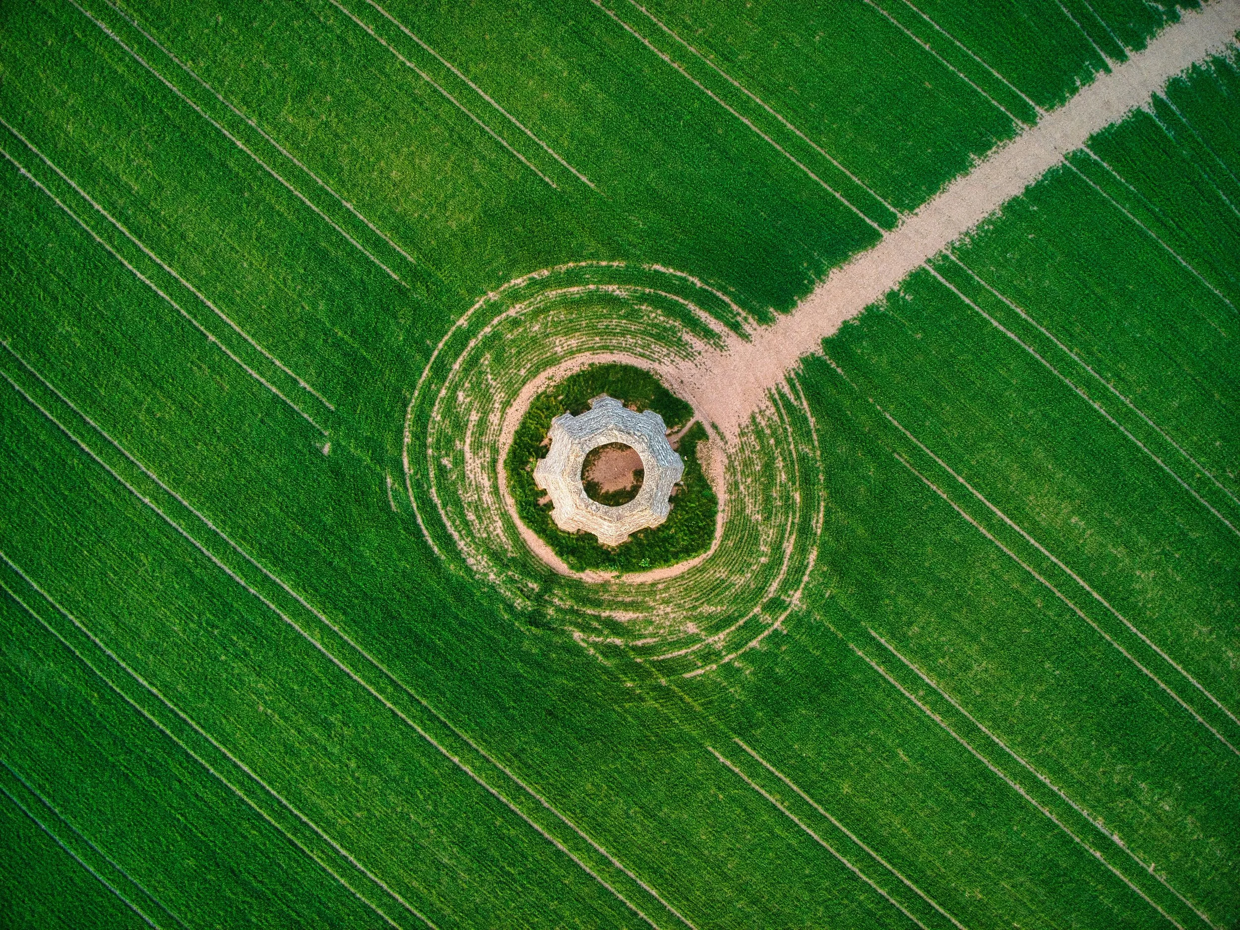 An aerial view of a round stone structure with a circular opening in the center, surrounded by green fields with parallel lines, and a dirt path leading to the structure.