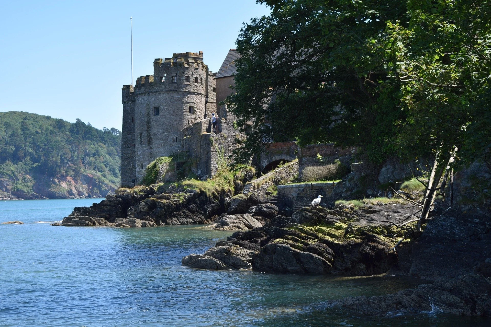 A stone castle on a rocky shoreline by a body of water, with trees in the foreground and hills in the background.