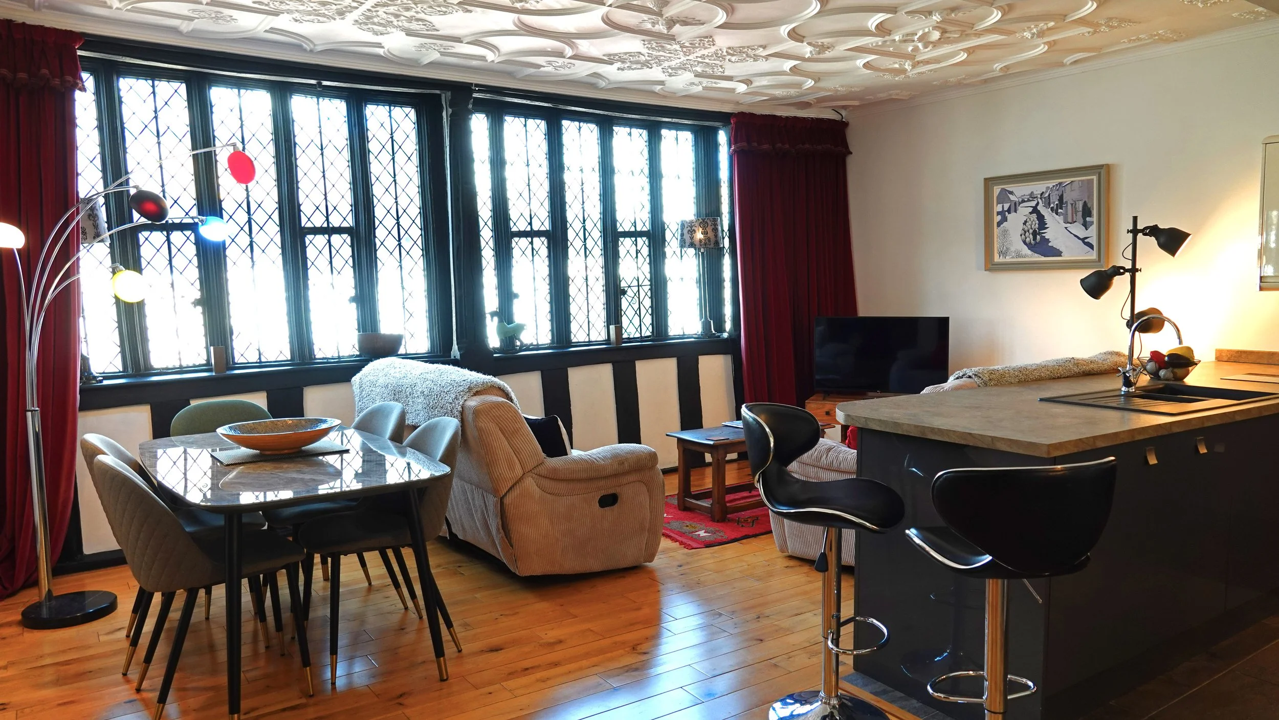 Living room with large stained glass windows, a dining table with six chairs, a beige recliner, a sofa, a TV, and a kitchen with black bar stools and a beige countertop.
