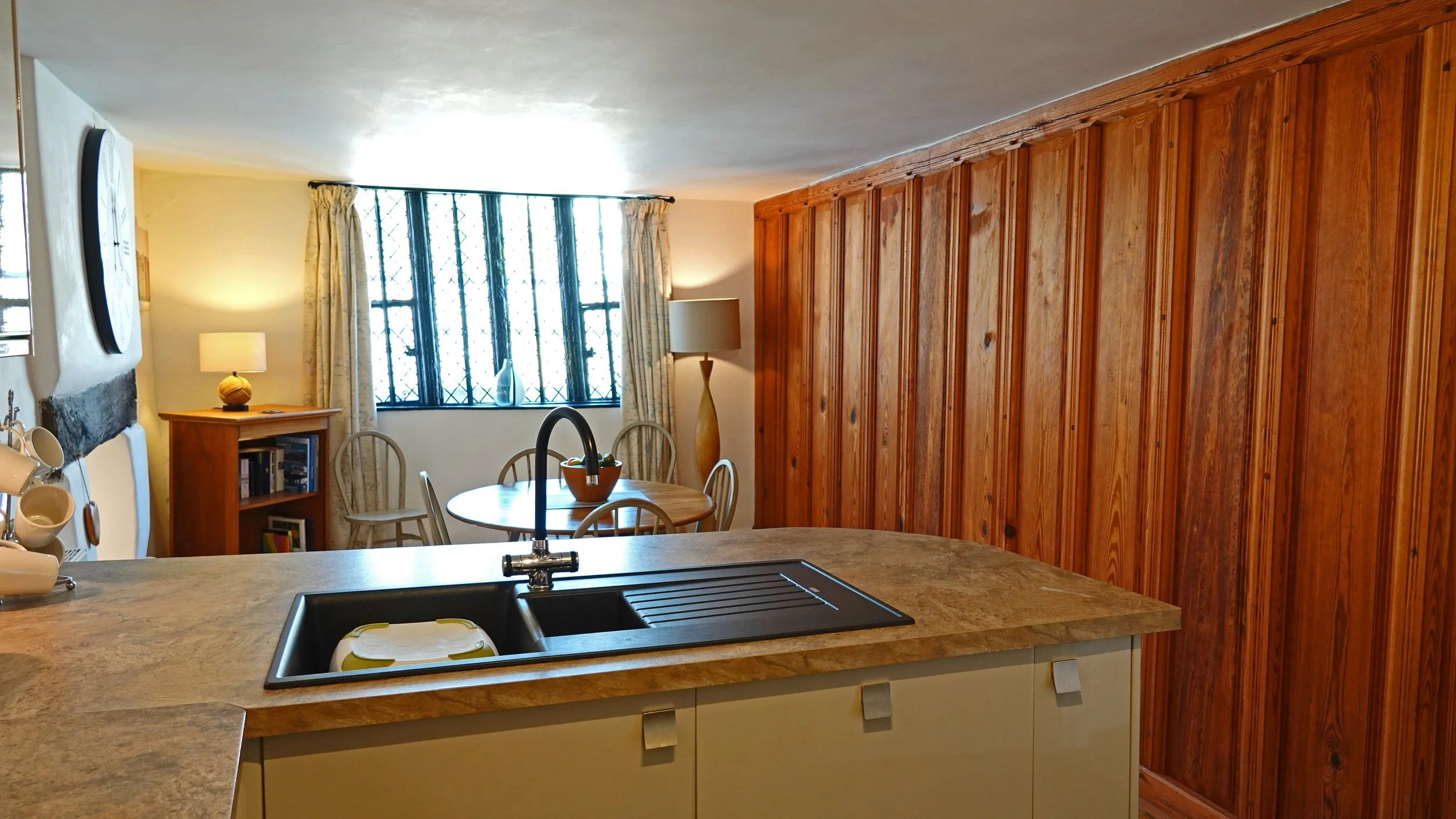 Kitchen with granite countertop and a black sink, wooden cabinets, a dining round table with chairs, a window with black metal bars and curtains, a floor lamp, a bookshelf, and a small lamp on a wooden cabinet.