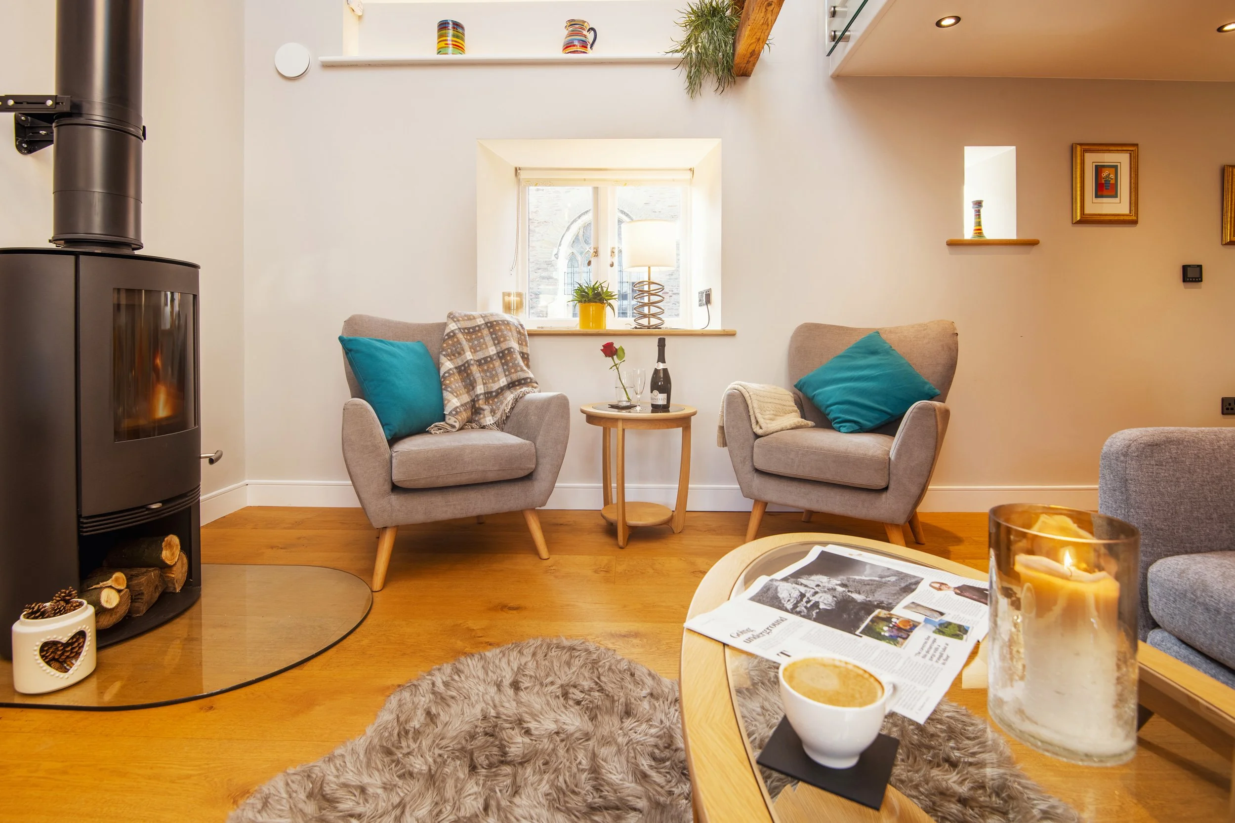 A cozy living room with two beige armchairs, each with a blue cushion, positioned near a small window with a yellow vase, plants, and a lamp. There's a black wood-burning stove on the left with logs underneath, a shaggy rug on a wooden floor, and a round wooden table with a bottle of wine, a rose, and glasses. A coffee table in the foreground holds a newspaper, a candle, and a coffee cup.