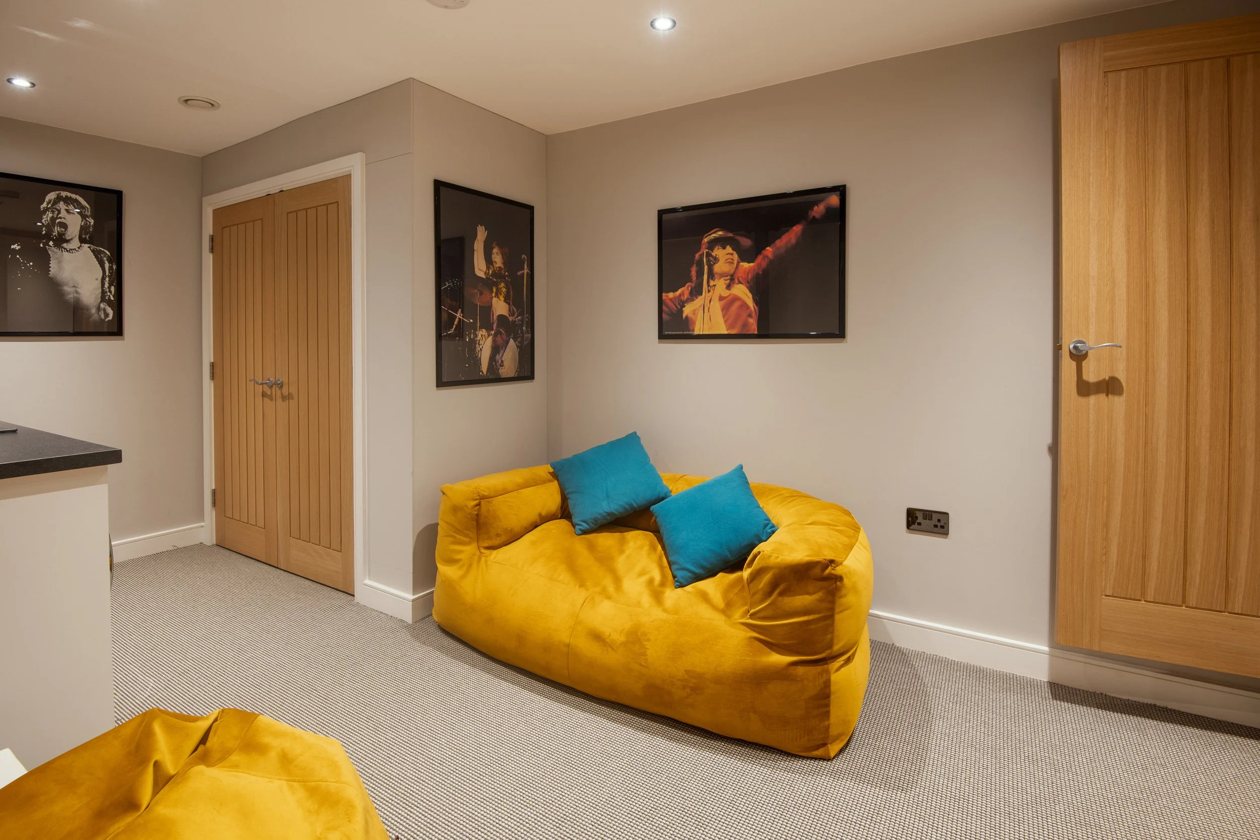 A living room with a yellow velvet sofa, two blue cushions, framed wall art including a black and white photo and colorful stage photos, wooden doors, and a beige carpet.