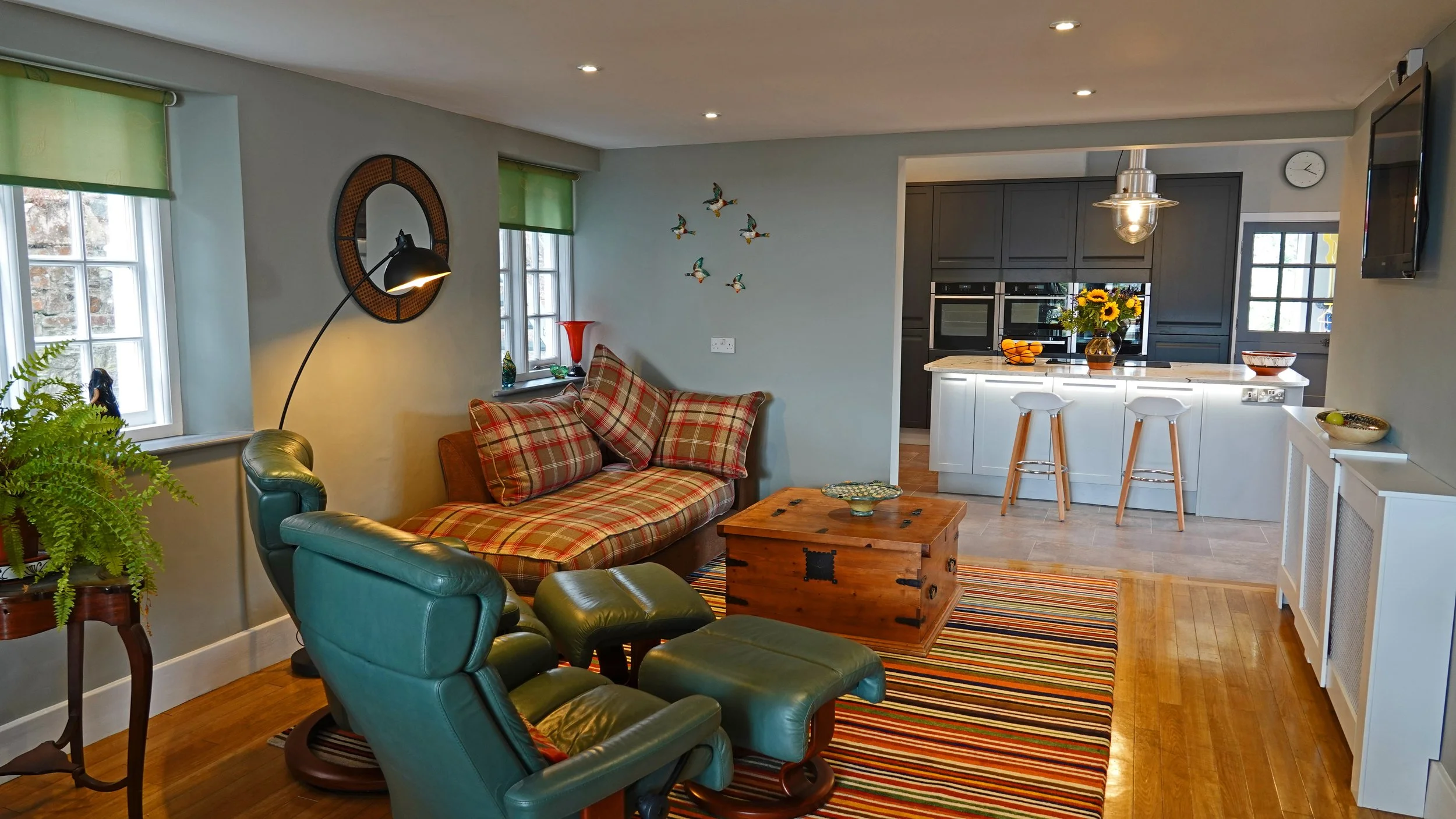 Living room with green leather chairs, a plaid couch with matching pillows, a wooden chest coffee table, striped area rug, and a kitchen with white island, gray cabinets, sunflower bouquet, and bar stools.