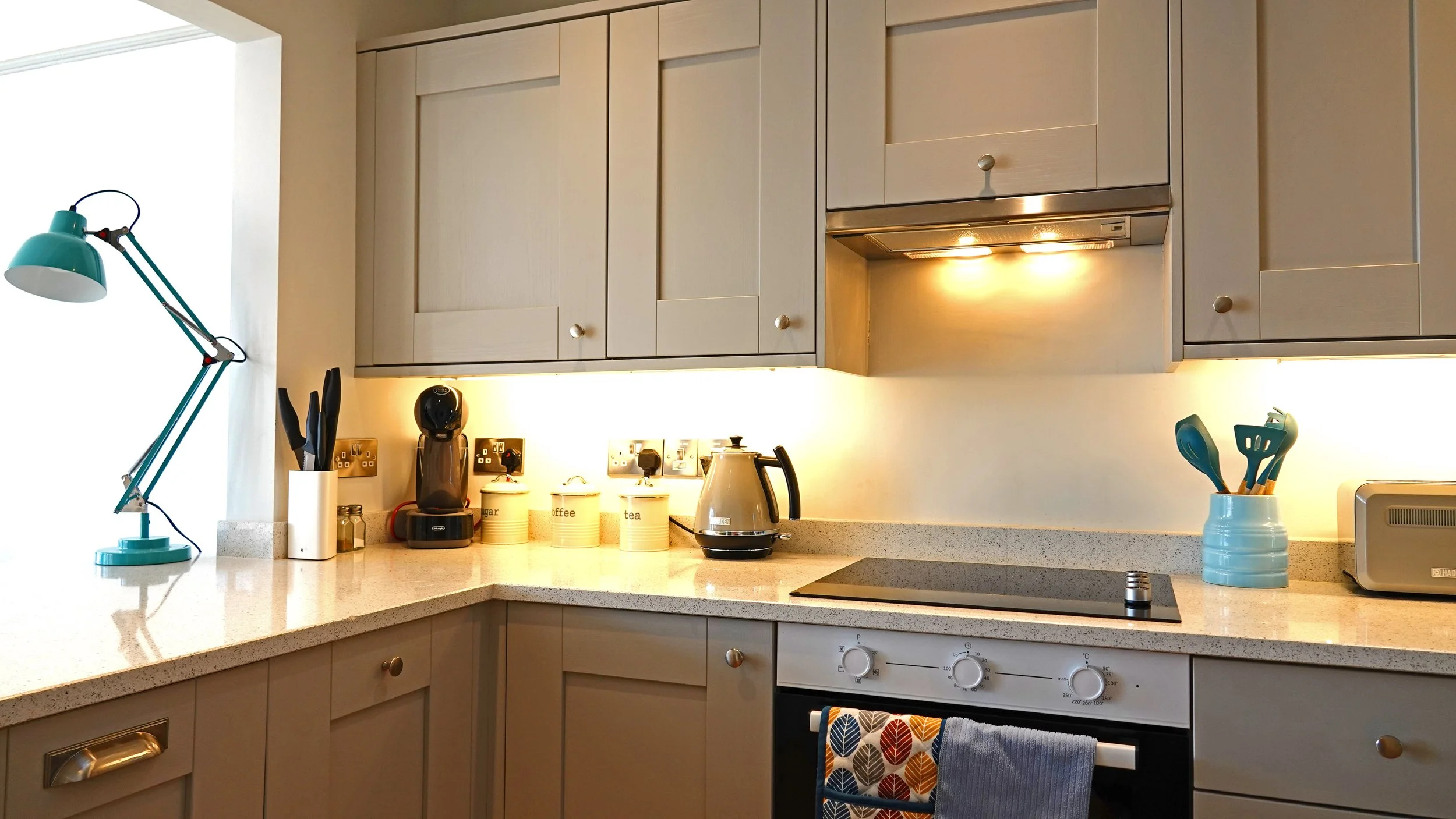 Kitchen countertop with a blue desk lamp, knife holder, coffee machine, labeled jars for sugar, coffee, and tea, electric kettle, stovetop, utensils in a blue mug, toaster, and beige cabinets.