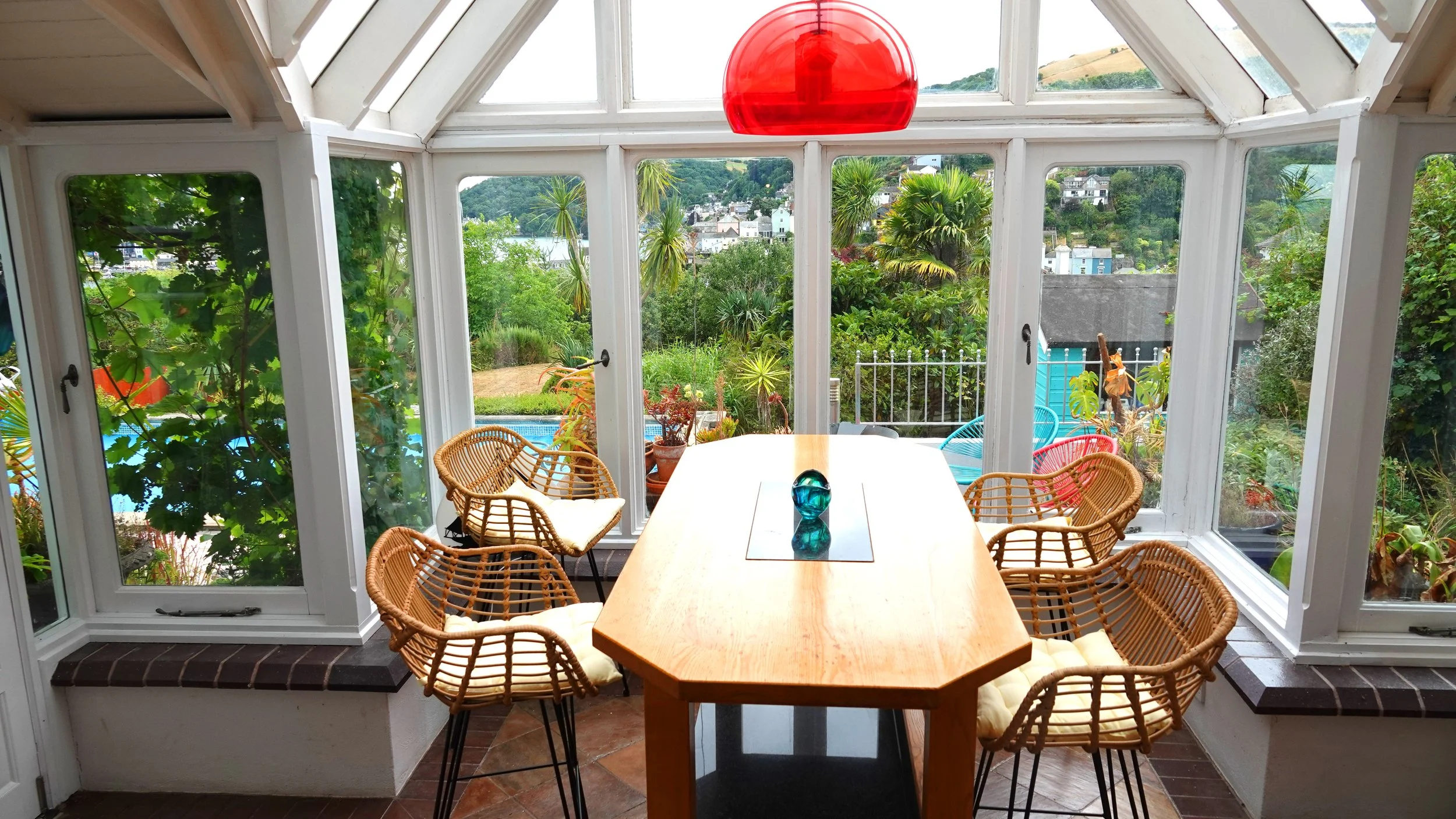 Sunroom with a wooden dining table, wicker chairs with cushions, large windows showing a garden, trees, and hills, and a red hanging lamp