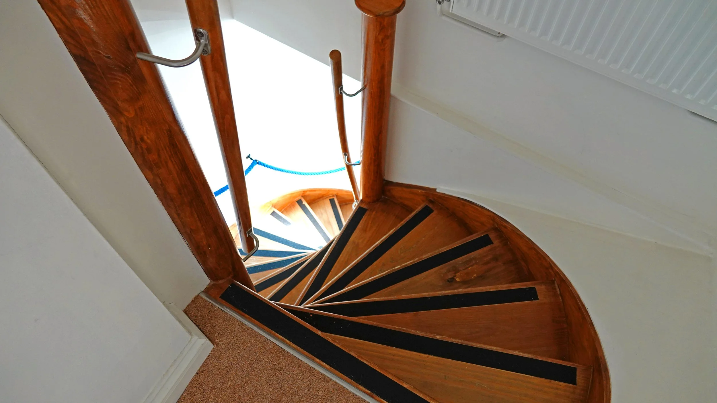 Top-down view of a spiral staircase with wooden steps and black anti-slip strips, with a wooden handrail on a white wall.