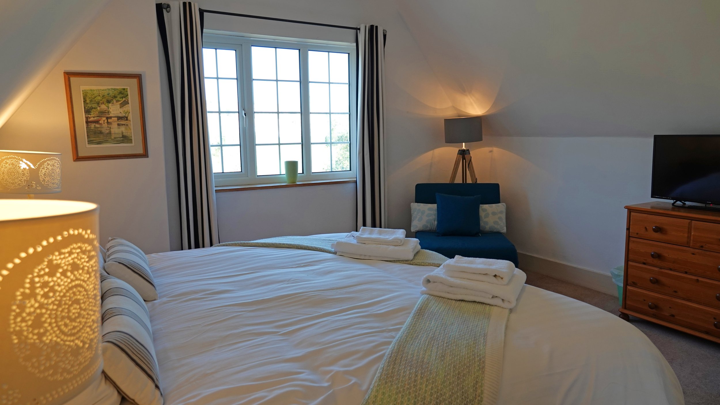 A cozy bedroom with a large bed covered in white sheets and striped pillows. Towels are folded on the bed. There is a window with black and white curtains, a framed picture on the wall, and a small sitting area with a blue chair and pillows. A wooden dresser with a flat-screen TV sits next to a floor lamp, and sunlight filters into the room.