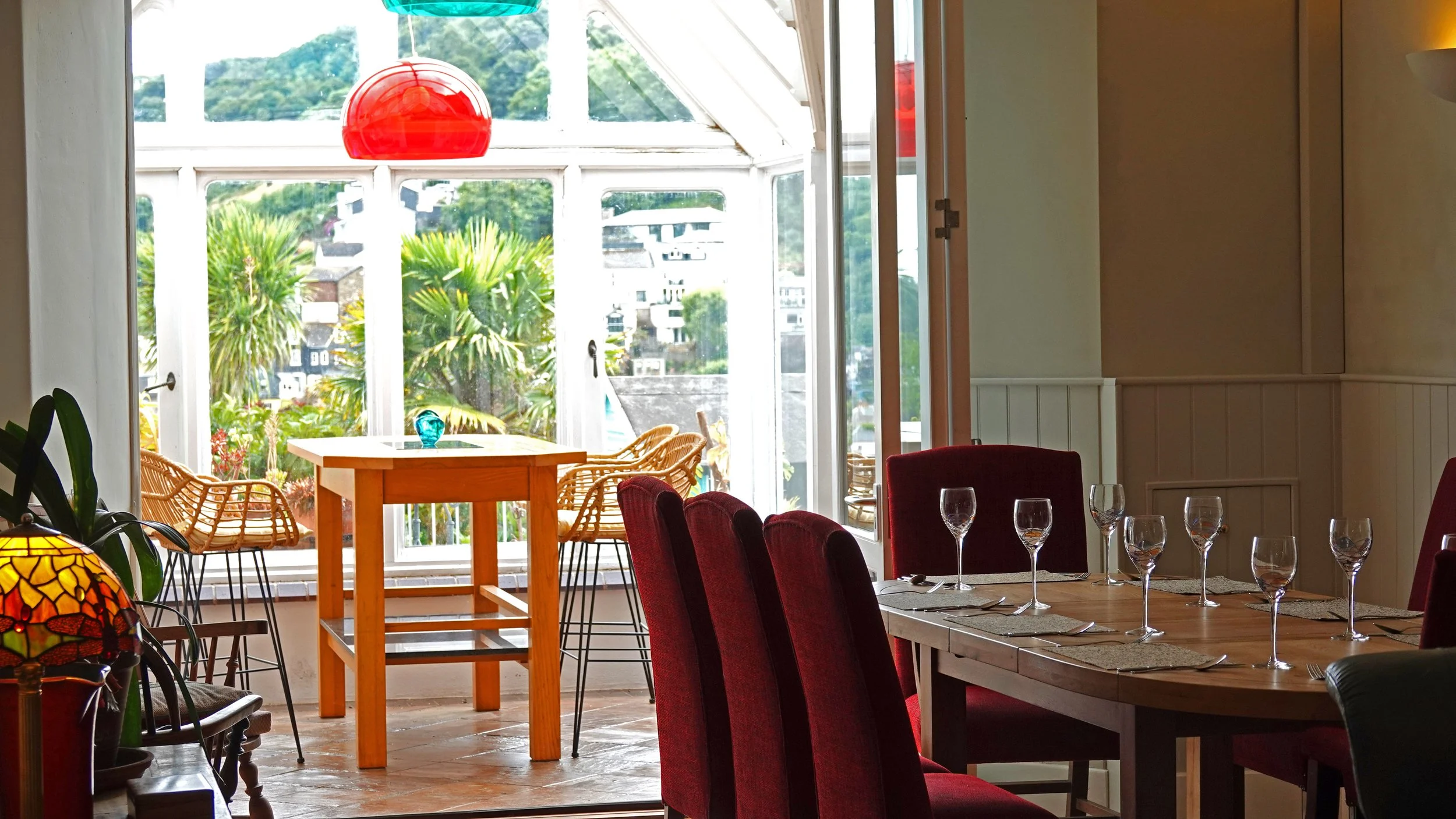 Interior view of a dining area with a large wooden table set with wine glasses and napkins, surrounded by red velvet chairs, with a glass ceiling and open doors revealing a patio area with wicker chairs and greenery outside.