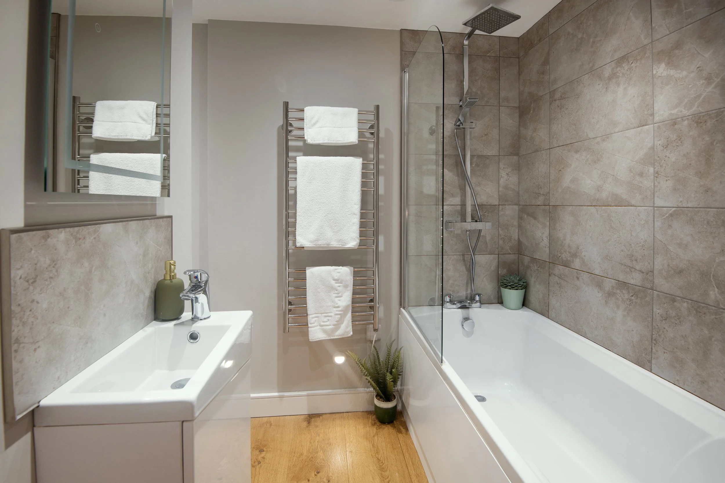 Modern bathroom with a bathtub, shower, small sink, towel rack, mirror, and potted plants.